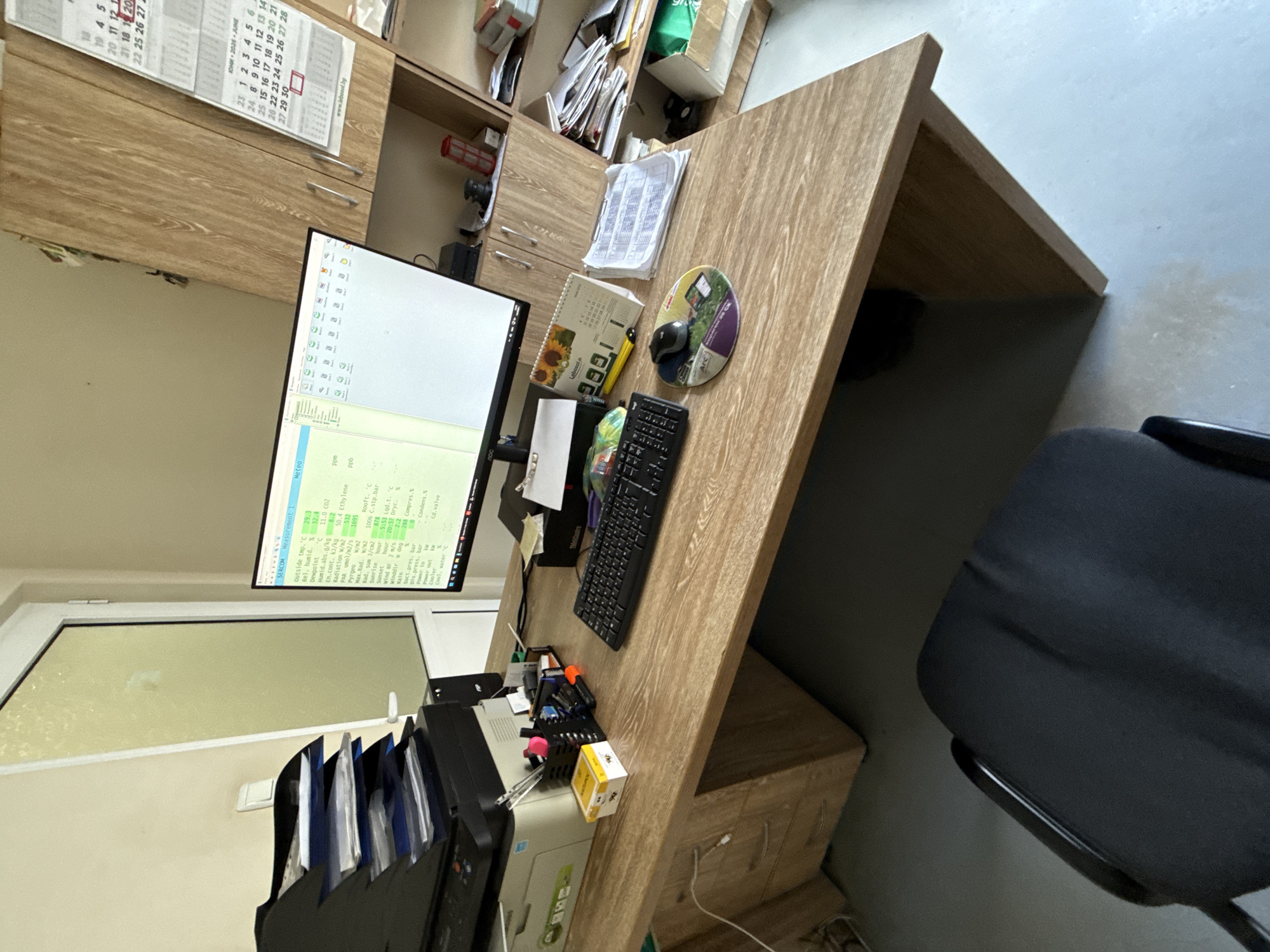 This is a medium-shot, high-angle photograph taken indoors, featuring a cluttered workspace at an office-type setting.
The primary focus is a desk with a monitor displaying code or data. The desk also holds a keyboard, mouse on a mousepad with images, a calendar, papers, and office supplies including pens and a container of small yellow cubes. A printer is visible on the left side of the desk.
The room appears to have a window, which is slightly open, with a white frame. The walls are light-colored.
To the right of the desk is a black office chair, partially visible.
Behind the desk are wooden cabinets and shelves filled with various items, including papers and documents.
The scene is not explicitly identifiable as Stara Zagora, Bulgaria, and does not include any people.