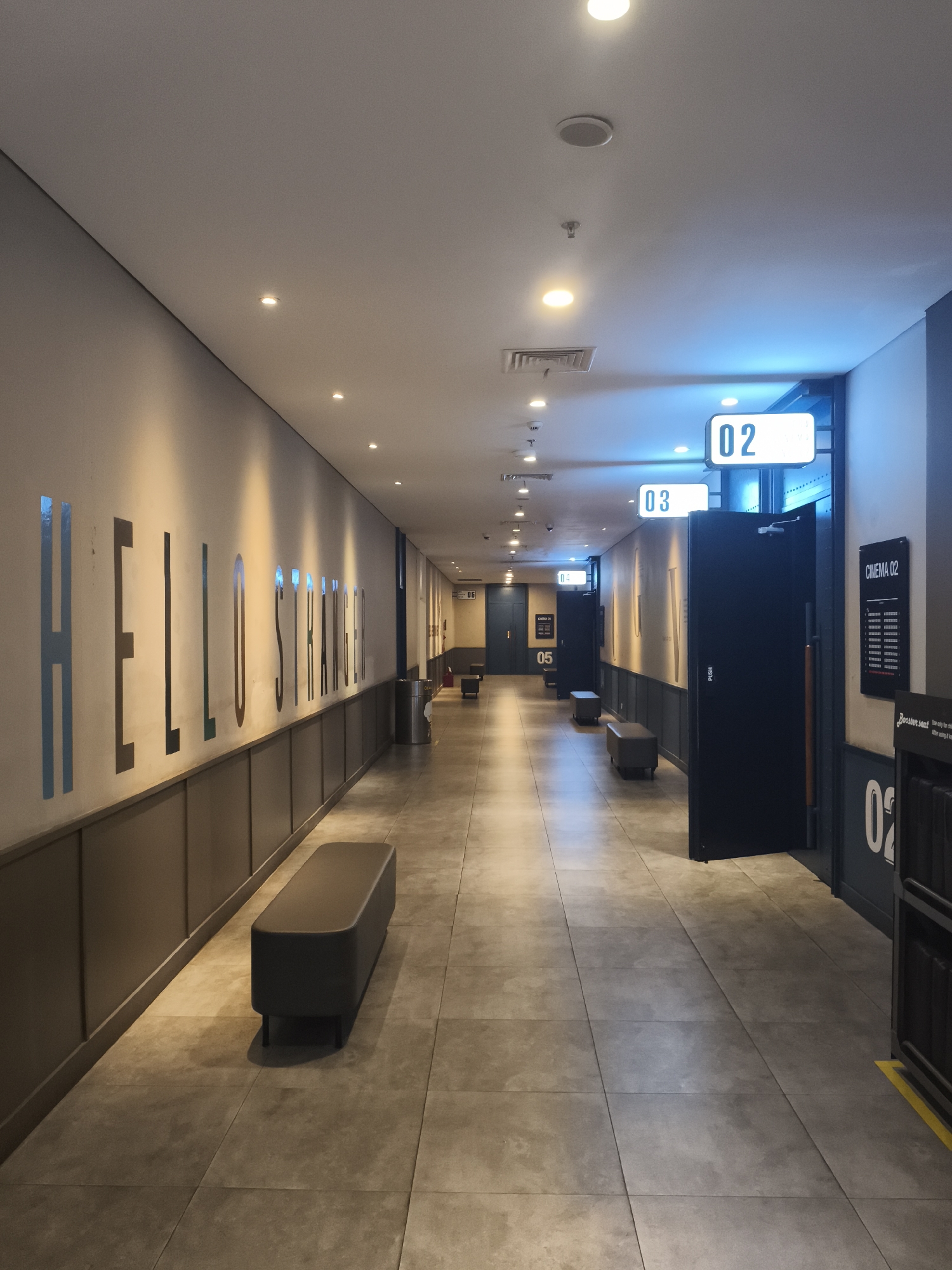 The image shows a long, empty hallway at a cinema in Depok, Indonesia. The hallway is lined with doors on the right, marked with numbers "02", "03", "04", "05", and "06", indicating cinema halls. The walls are adorned with large, cut-out letters that spell "HELLO STRANGER" on the left, illuminated by warm lighting. The floor is tiled with large, grey squares. There are several dark grey benches placed along the left wall. The ceiling features a series of recessed lights. The overall atmosphere is clean and modern, typical of a contemporary cinema entrance or waiting area. No people are visible in the image.