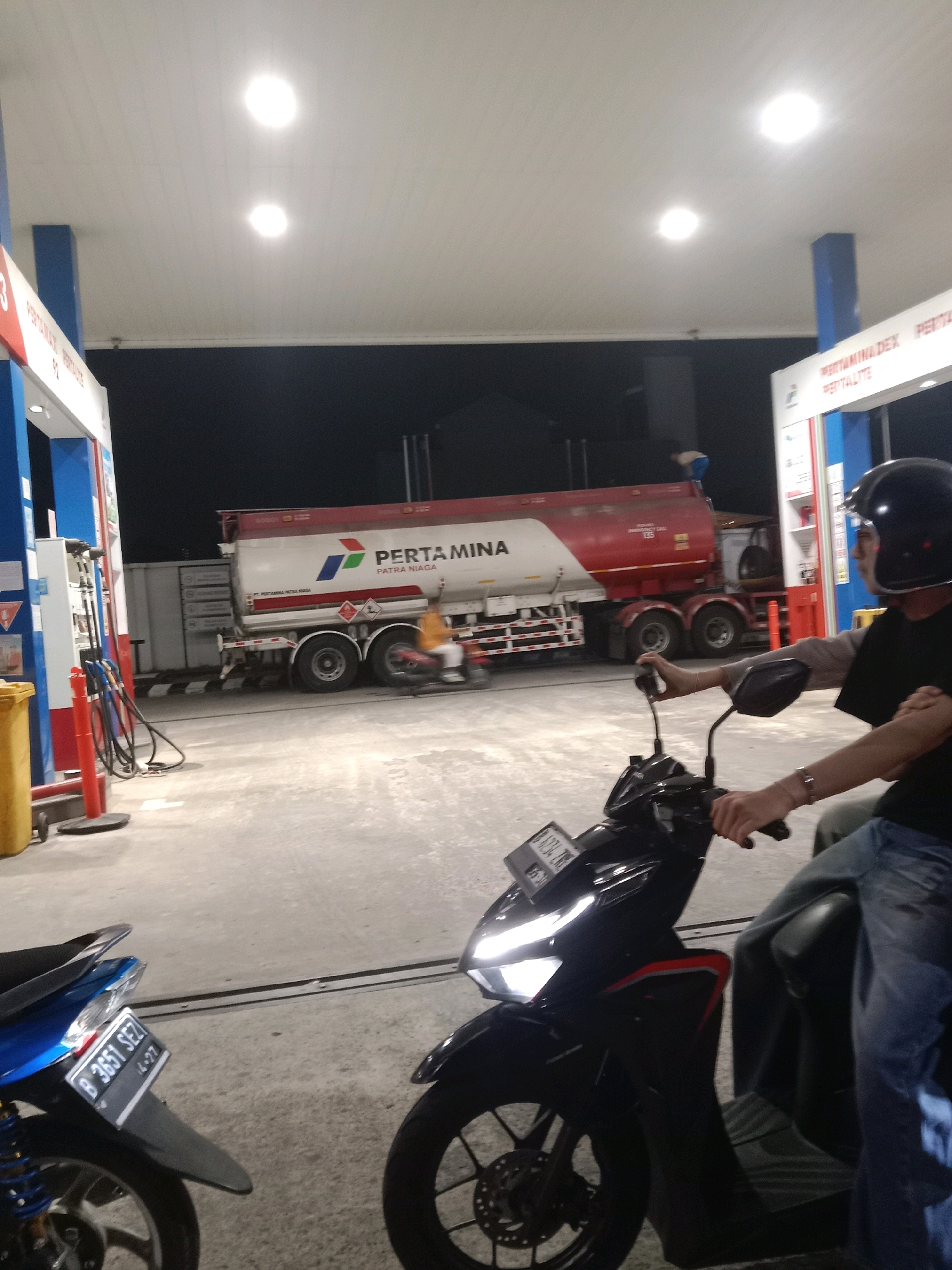 The image depicts a scene at a Pertamina gas station in South Tangerang, Indonesia, at night. In the foreground, two motorcycles are parked. The motorcycle on the right has two people on it, with the rider wearing a black helmet and the passenger sitting behind. The motorcycle on the left is partially visible and is blue. In the background, a large red and white Pertamina tanker truck is parked, with its logo prominently displayed. The gas station appears to be well-lit with overhead lights. The ground is paved concrete. The overall atmosphere is that of a busy, operational gas station at night.