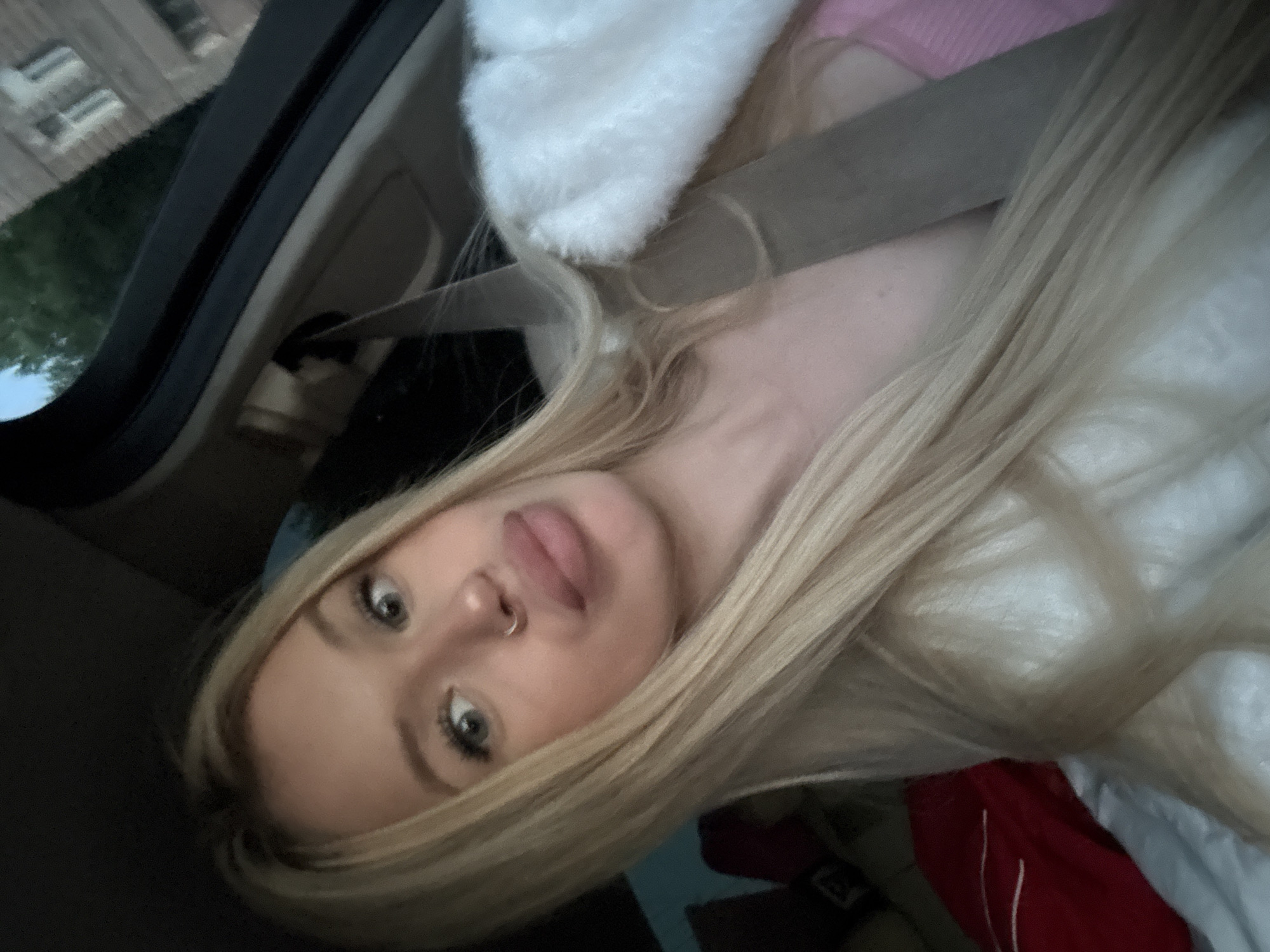 The image shows a close-up, vertically oriented selfie of a young blonde woman inside a vehicle. She is wearing a seatbelt over a light-colored, fluffy garment. Her hair is long and blonde, falling around her face. She has a nose ring and is looking towards the camera with a neutral expression. The background suggests the interior of a car, with a seatbelt visible and dark interior paneling. Outside the car window, trees and a building with windows can be seen, hinting at an outdoor setting, possibly in an urban or suburban area. The lighting appears to be natural, suggesting it is daytime. The overall mood is casual and personal. There is no visible text in the image.