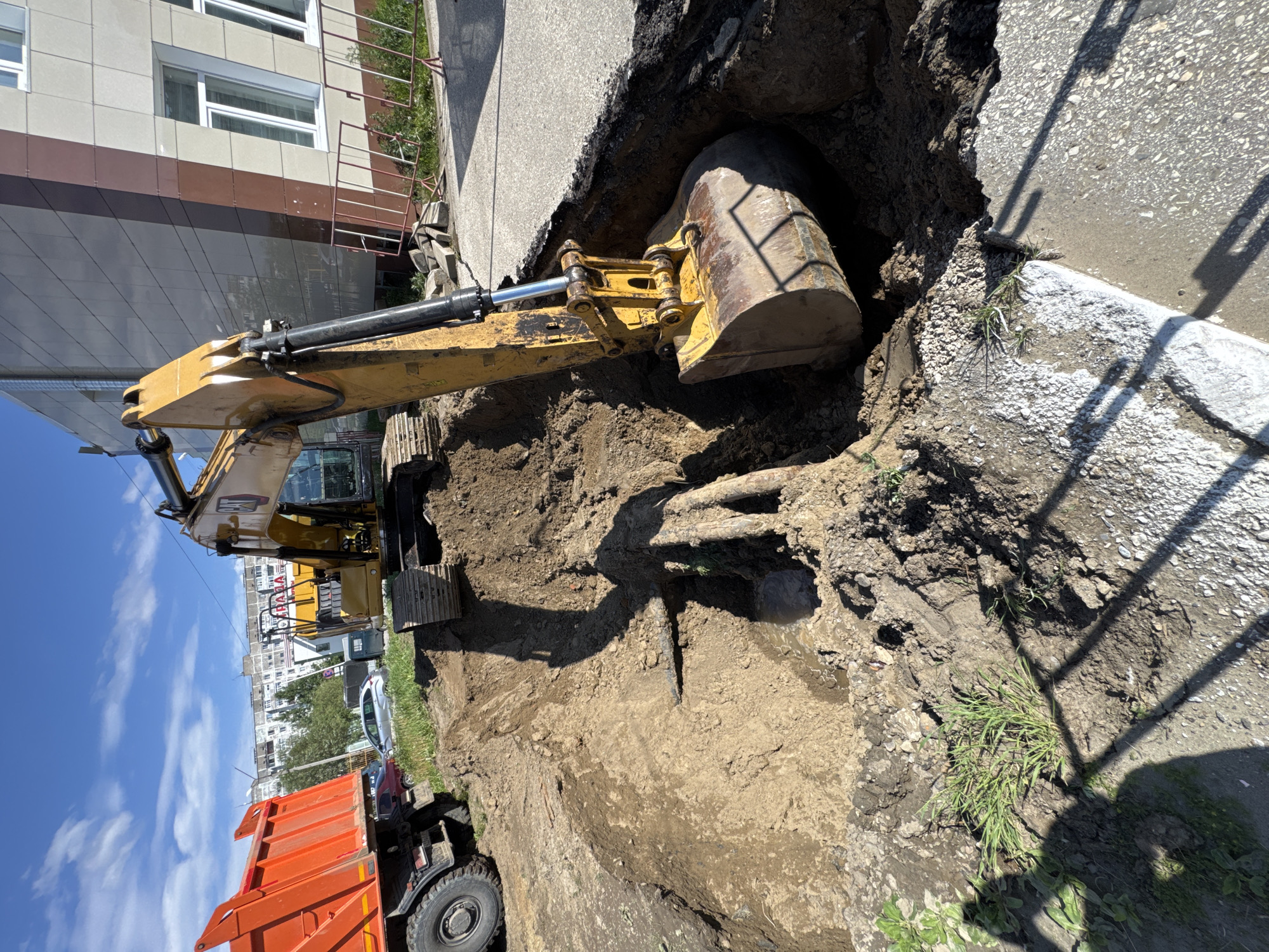 An excavator is digging a trench in the ground next to an orange dump truck. The trench is deep and filled with dirt and some water. There are pipes visible in the trench. In the background, there is a building with modern architecture, a metal fence, and some trees. The sky is blue with some clouds. The scene appears to be a construction site.