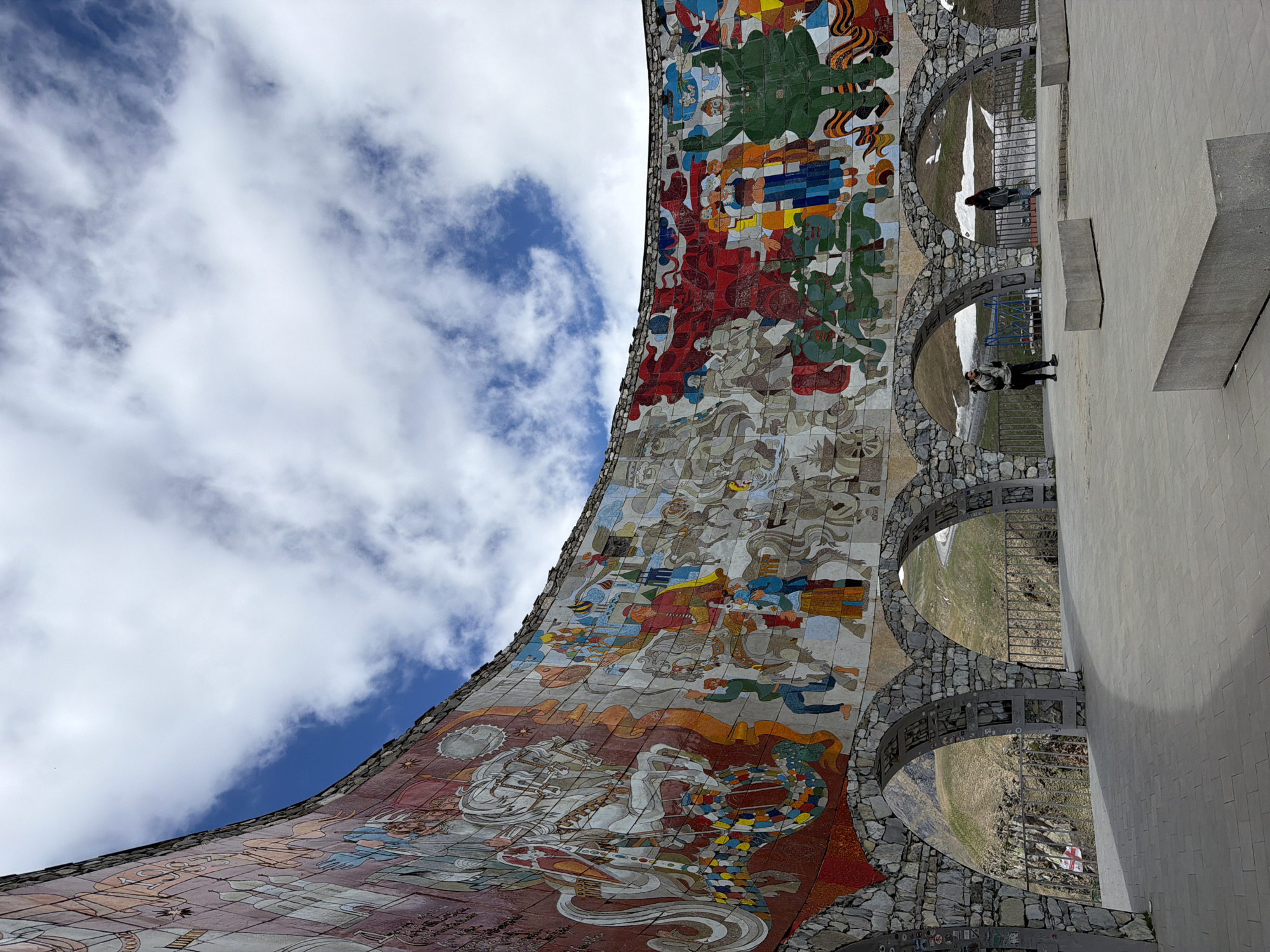The image shows a section of the Georgia–Russia Friendship Monument located in Gudauri, Georgia. The monument is a large concrete and mosaic structure. A curved mosaic mural features historical and cultural scenes. Two people are visible on a walkway with arches alongside the mural. The sky behind the mural is blue with white clouds. A white concrete wall is visible on the right side of the image. A landscape is visible through the arches of the walkway. The image is taken from a low angle, looking upwards.