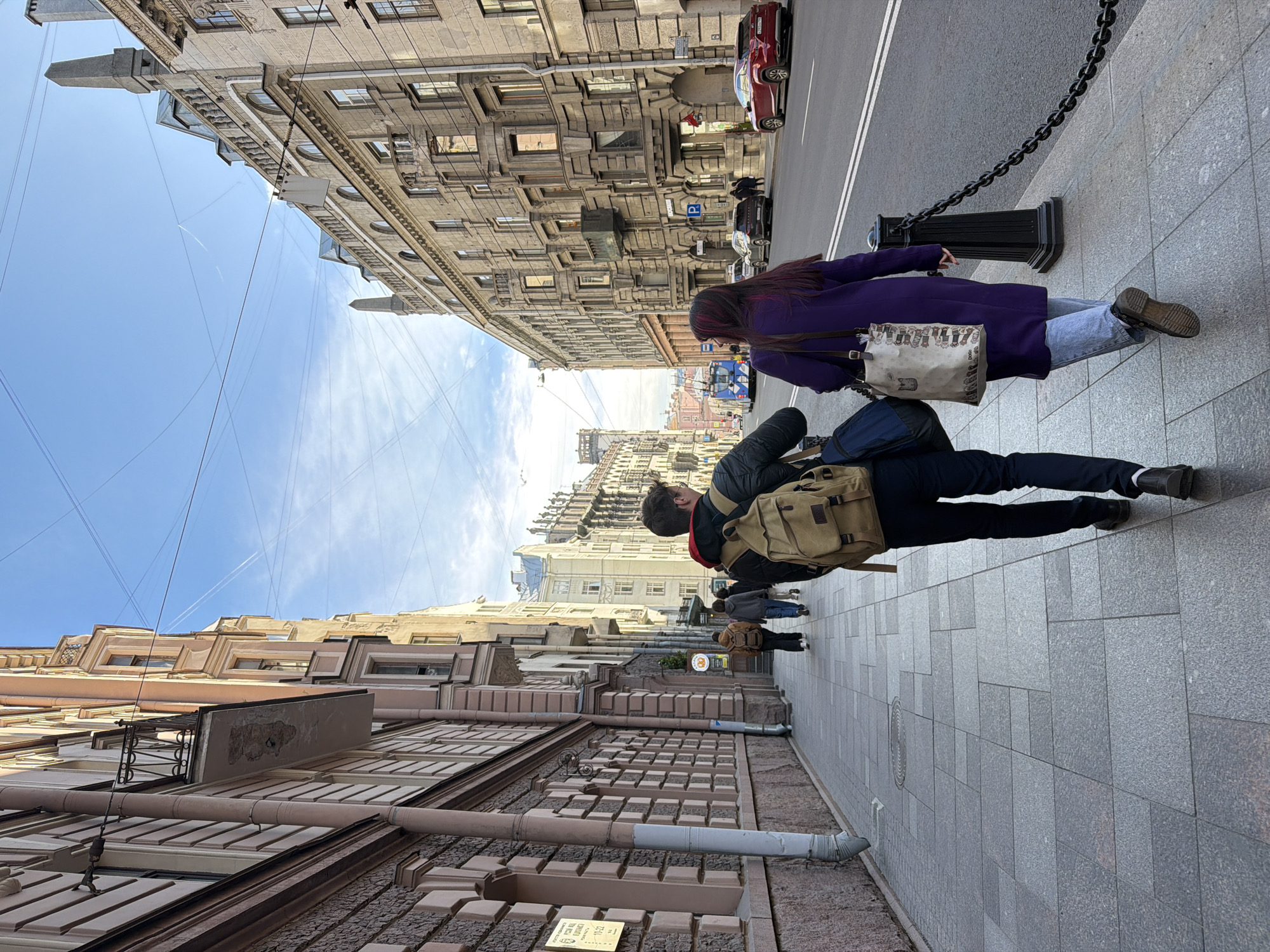 A street scene photographed in Saint Petersburg, Russia, shows two people walking on a sidewalk. A woman in a purple coat and jeans carries a patterned tote bag. A person with a tan backpack walks beside her. The street features cars and architectural buildings. Overhead, power lines crisscross against a pale blue sky with faint clouds.
