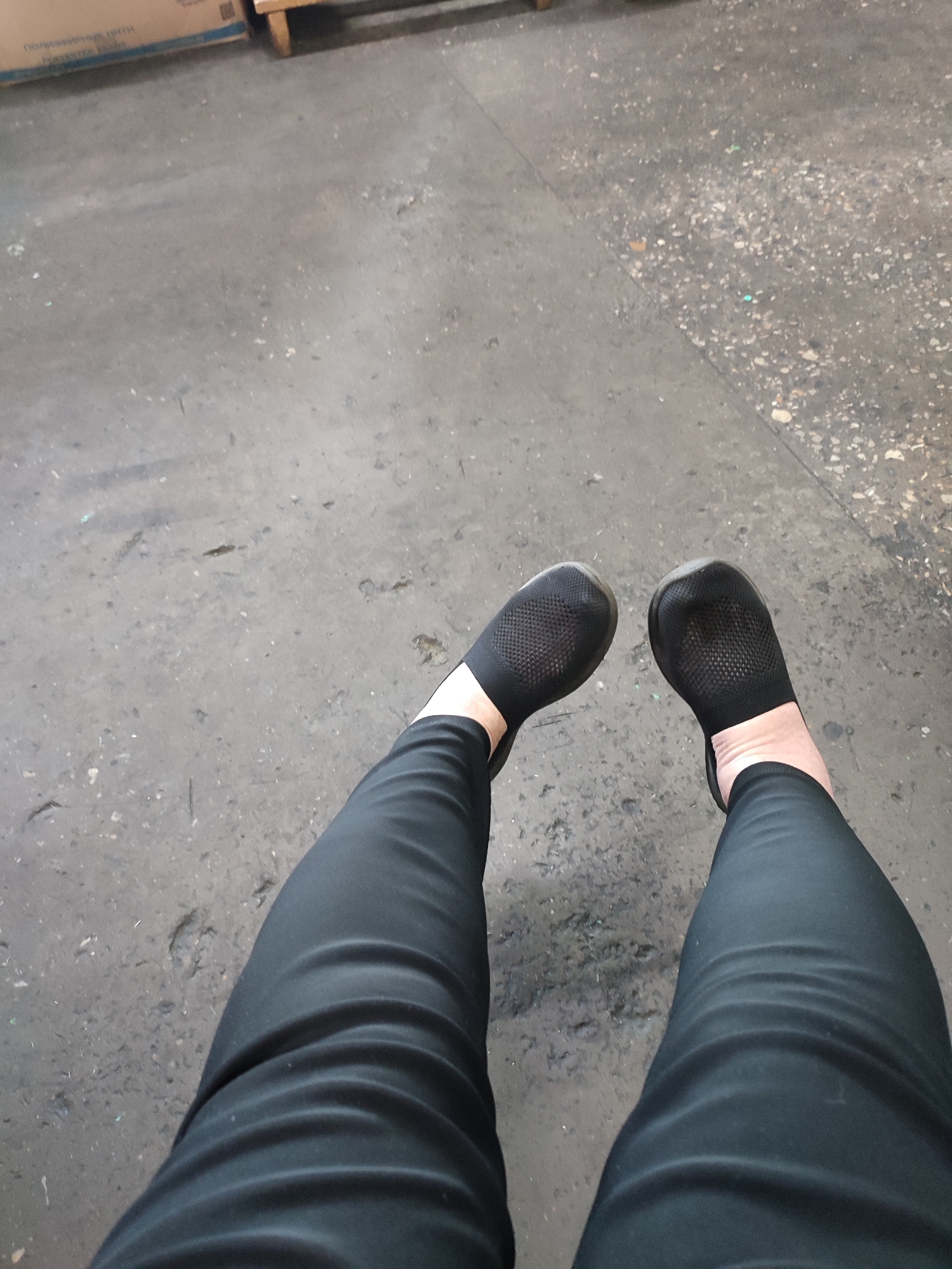 The image displays a pair of legs from the knees down, clad in black leggings and black slip-on shoes. The legs are positioned on a rough, concrete-like surface, appearing to be outdoors. The background is a textured gray ground with scattered debris. To the upper left of the image, a portion of a cardboard box is visible. The overall scene suggests an urban or industrial setting. There are no people or animals clearly identifiable beyond the subject whose legs are depicted. The lighting is even, indicating it is daytime. The image appears to have been taken from a slightly elevated perspective, looking down at the subject's legs. There are no specific landmarks or elements that definitively place the location in Narowlya, Belarus, as the environment is generic.