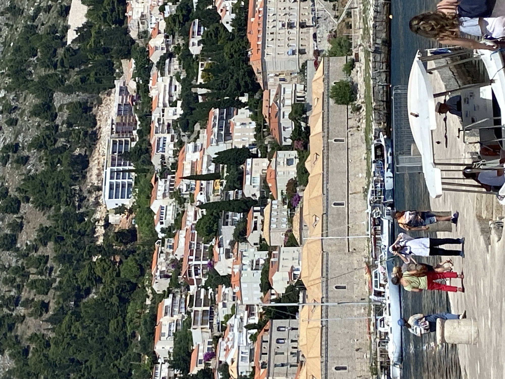 The image depicts a scenic view of Dubrovnik, Croatia, characterized by its iconic terracotta-roofed buildings cascading down a hillside towards a harbor. In the foreground, several people are gathered on a stone pier. A woman with a backpack walks away from the viewer, while a group of three individuals stand together, one adjusting their hat. To the right, a small white boat with a canopy, likely a water taxi or tour boat, is docked. The boat has a driver at the helm. Behind the pier, several larger boats and yachts are moored in the clear blue water. The middle ground is dominated by a long, stone building with arched windows, likely part of the city's historical fortifications or waterfront structures. Above this building, the hillside is densely covered with lush green trees, and a multitude of white and cream-colored houses with red tile roofs are built into the slope. Further up the hill, a modern white building with multiple balconies stands out. The overall scene captures the vibrant atmosphere of Dubrovnik's waterfront, blending historical architecture with the natural beauty of its coastal setting.