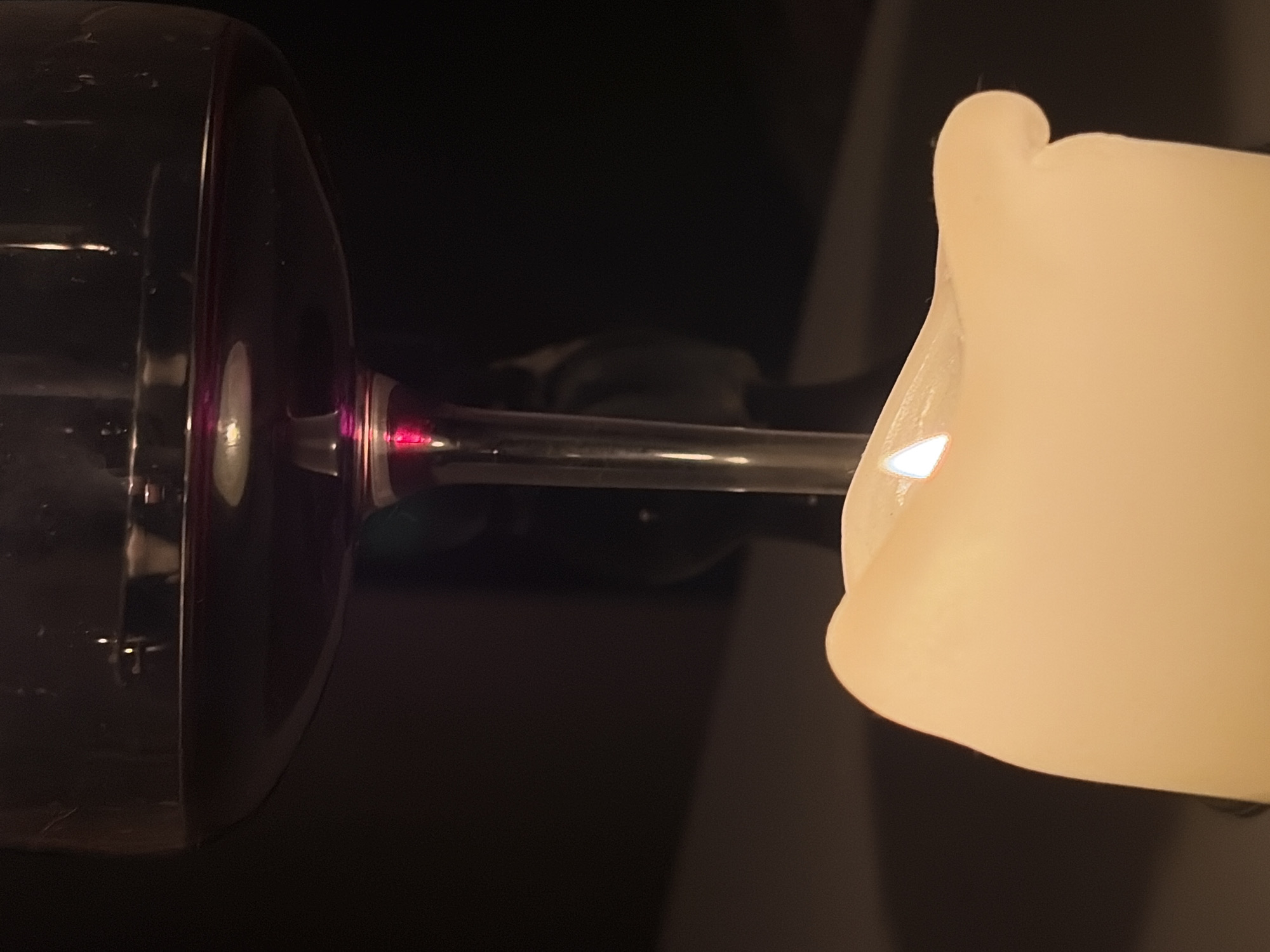 The image depicts a wine glass with a partially visible background. The wine glass has a large bowl and a slender stem, appearing to be illuminated by a light source from the right. The bowl of the glass is mostly in shadow, but has a dark purple rim. The background is blurred, creating a sense of depth. Based on the image, it is not possible to determine the location in Budva, Montenegro.
