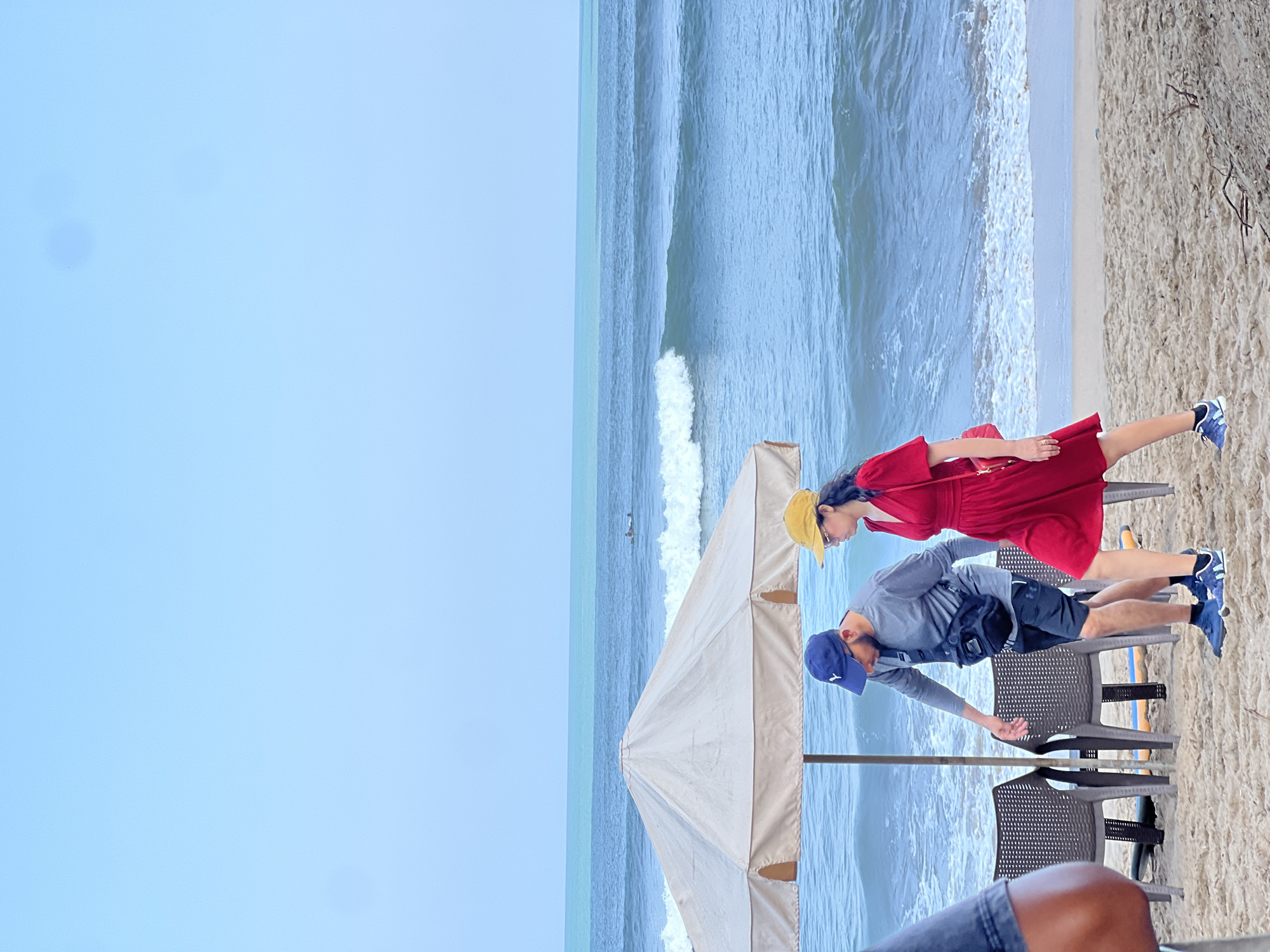 The image depicts a beach scene in Kuta, Indonesia, with two people walking on the sand. A man wearing a gray long-sleeved shirt, black shorts, a blue baseball cap, and a black fanny pack is walking towards the left. He is wearing blue athletic shoes. A woman wearing a red dress, a yellow hat, and carrying a red purse is walking towards the right, beside the man. Both are walking in front of a large, light-colored beach umbrella. Behind them, the ocean displays blue water with white waves breaking near the shore. The sky is a clear, pale blue, and the beach is covered in light brown sand with some scattered debris. Two empty chairs are visible under the umbrella.