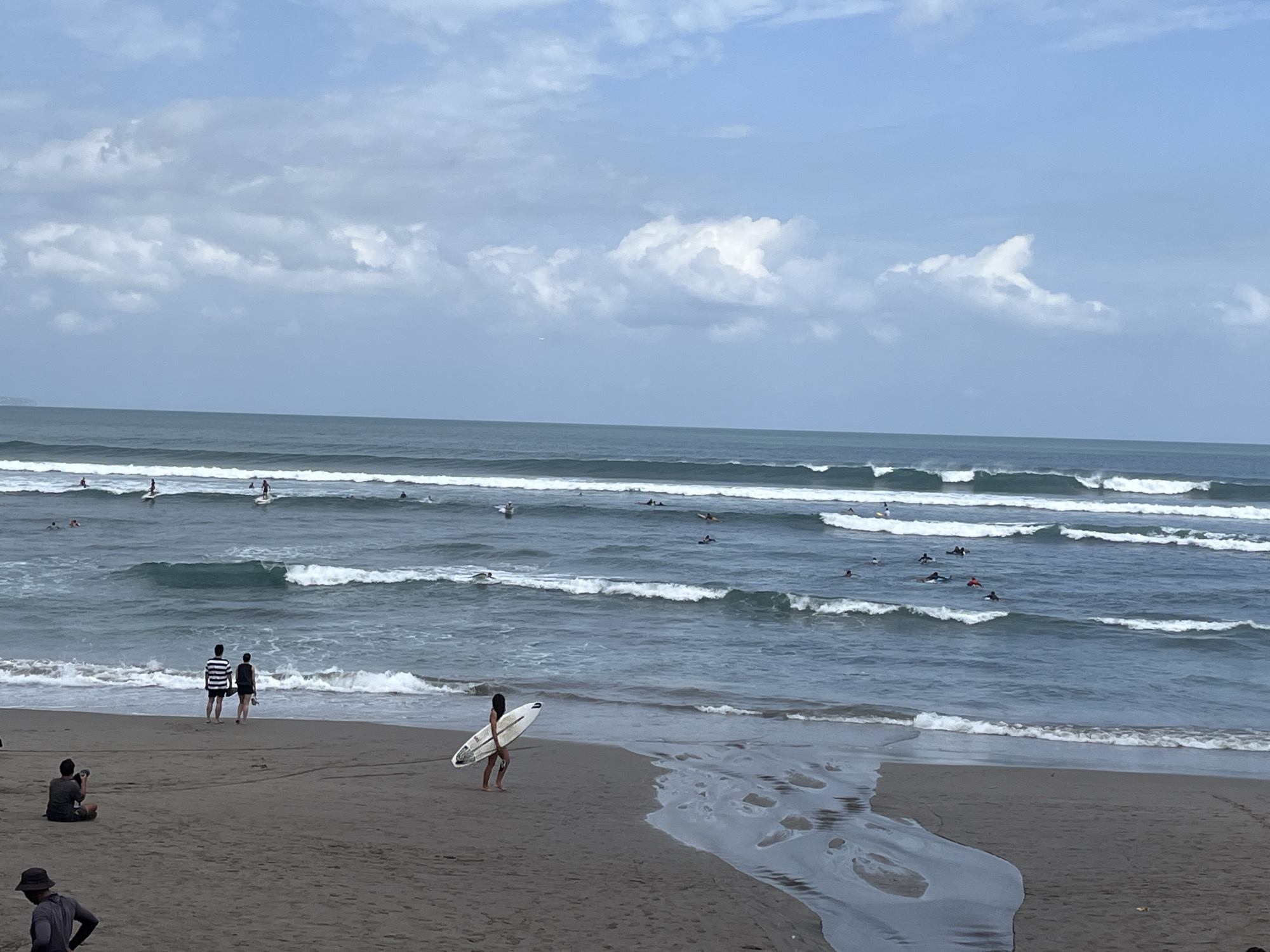 An eye-level, medium shot captures a beach scene in North Kuta, Indonesia, under a partly cloudy sky. The ocean is active with multiple sets of waves, and numerous surfers are visible in the water, some riding waves and others paddling. On the sandy beach in the foreground, a group of people are present. Two individuals stand near the water's edge, facing the ocean. To the left, a person is seated on the sand, holding a camera. Further to the right, a person walks along the shoreline, carrying a surfboard. A small stream of water flows from the beach into the ocean on the right side of the frame. The sand is a dark, wet color, suggesting recent moisture.