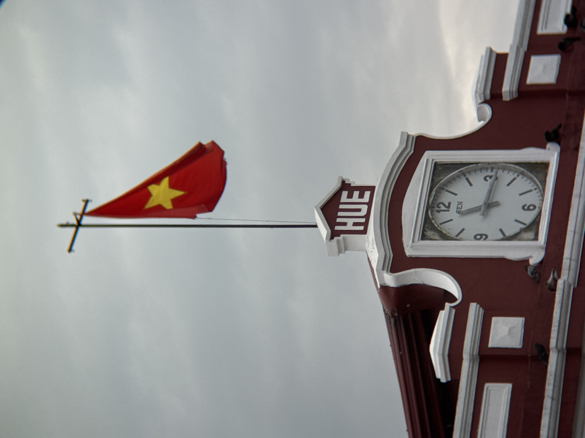 A Vietnam flag flies from a flagpole atop a building. The flag is red with a yellow star in the center. The building is painted dark red with white trim. A clock is embedded in the building's facade, showing the time as approximately 4:05. Above the clock, the word "HUE" is visible in white letters. Several pigeons are perched on the building's ledge. The sky is overcast with gray clouds.