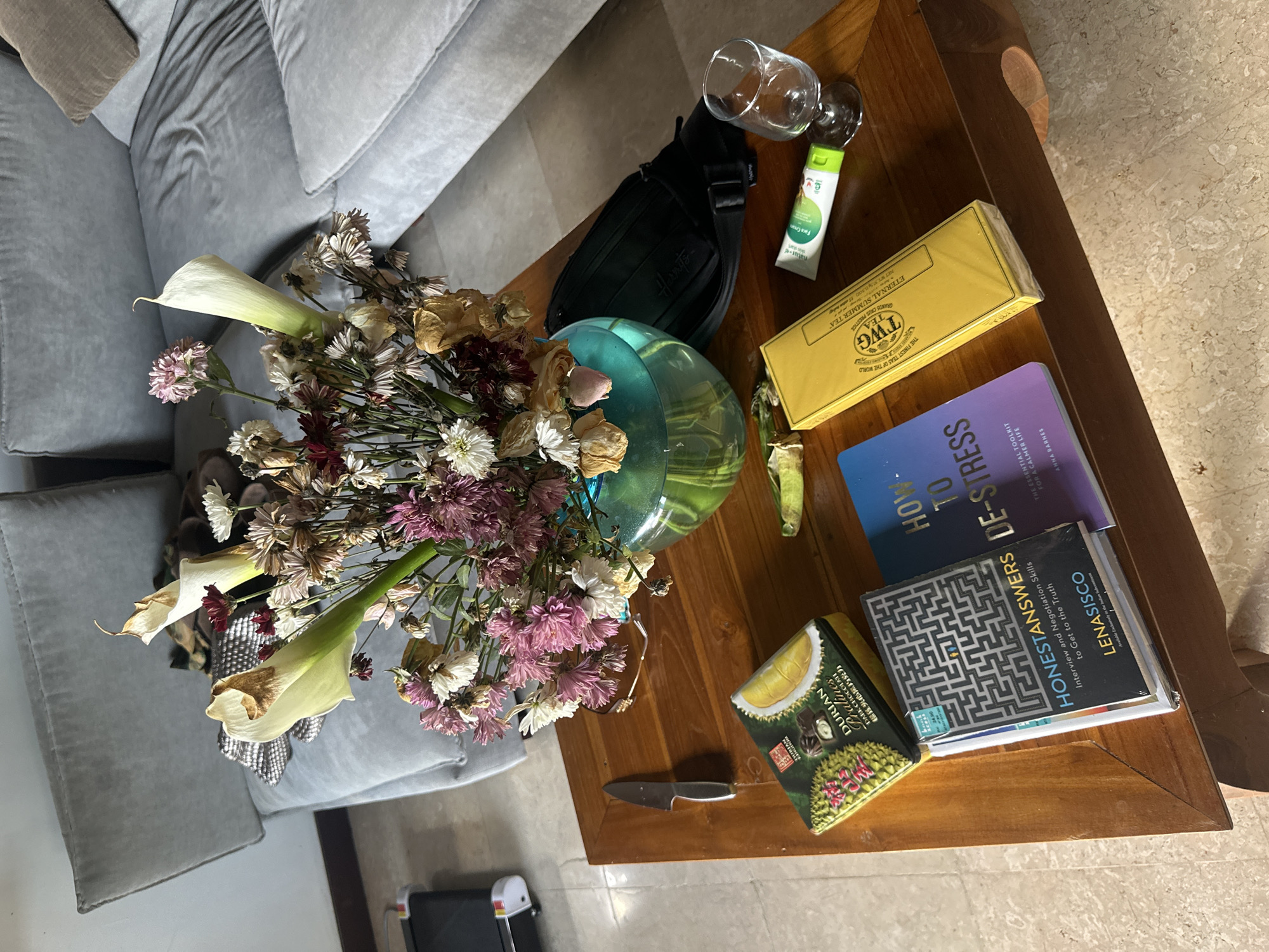 This overhead shot depicts a wooden coffee table in an indoor setting, likely a living room, with a light gray L-shaped sofa partially visible.
At the center of the table, a translucent teal glass vase holds a bouquet of flowers, many of which appear dried and wilted, including calla lilies and various types of chrysanthemums. The faded blooms suggest they have been there for some time.
Arranged on the table around the vase are several items. A black sling bag is seen near two empty clear glasses—one a wine glass and the other a tumbler. A small green tube labeled "Natur-e Face Cream" rests beside them. A yellow box of "TWG TEA ETERNAL SUMMER TEA GRANDS CRUS PRESTIGE THE FINEST TEAS OF THE WORLD" is also present.
Further right, a stack of two books includes "HOW TO DE-STRESS: THE ESSENTIAL TOOLKIT FOR A CALMER LIFE" by Anna Barnes and "HONESTANSWERS: Interview and Negotiation Skills to Get to the Truth" by Lenasisco, which features a maze pattern on its cover. A small box of "DURIAN Pralines DARK CHOCOLATE 相互夹心黑巧克力" with a durian illustration sits next to the books, and a small knife with a silver blade and black handle is also on the table.
The scene is lit by natural daylight, indicating it is daytime. No active event is taking place; the image is a still life composition of everyday items. The setting is an indoor space, consistent with a home or lounge in Kuta Selatan, Indonesia. The dried flowers contribute a notable detail, hinting at time passing or a temporary display.