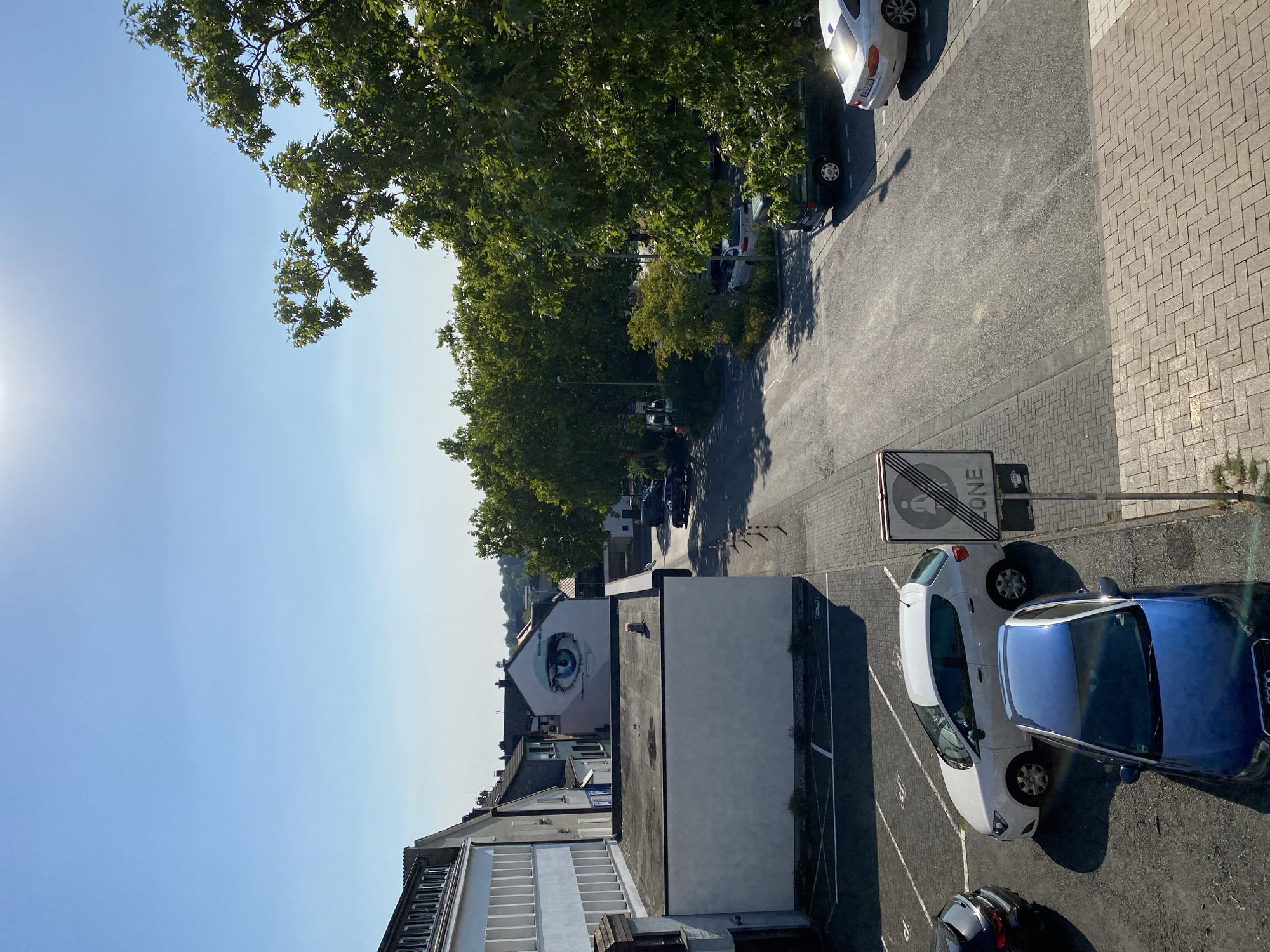The image depicts a view from an elevated position, looking down onto a parking area and street scene in Altenkirchen, Germany. The sky is clear and blue, with the sun visible and casting a bright glare.
In the foreground, a blue Audi A1 is parked. To its left, a white hatchback, possibly a Seat Ibiza, is parked in a marked spot. Further back, a dark-colored SUV is parked. On the right side of the image, a white BMW sedan is visible, parked on the street.
A street sign indicating a "ZONE" with a pedestrian symbol and a diagonal line is present. The street is paved with asphalt, and the sidewalk and parking area are paved with brick pavers in a herringbone pattern.
Buildings are visible on the left and in the background. The building on the immediate left is a modern structure with a white facade and rows of windows. Across the street, a building features a large mural of a blue eye. Other residential buildings with pitched roofs are visible in the distance, interspersed with mature trees. Green trees line the street and parking area, providing shade. The overall environment appears to be a mixed-use area with residential and commercial buildings.