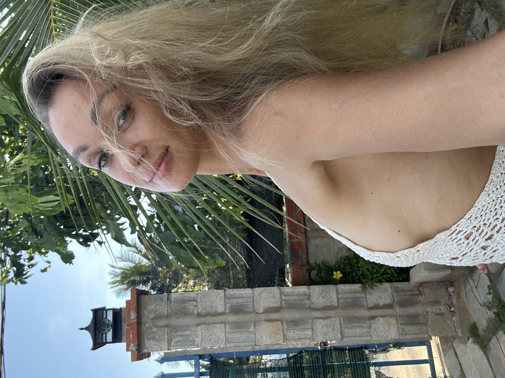 The image depicts a blonde woman taking a selfie outdoors. She is positioned with a slight tilt, her face close to the camera, with her hair cascading across the top of the frame. She wears a white, crocheted garment that reveals a portion of her shoulder and chest. The background includes a stone wall, a wrought-iron gate, various leafy trees and palm fronds, and a glimpse of the sky. The scene is consistent with a location in Mũi Né, Vietnam, based on the foliage and architectural elements.