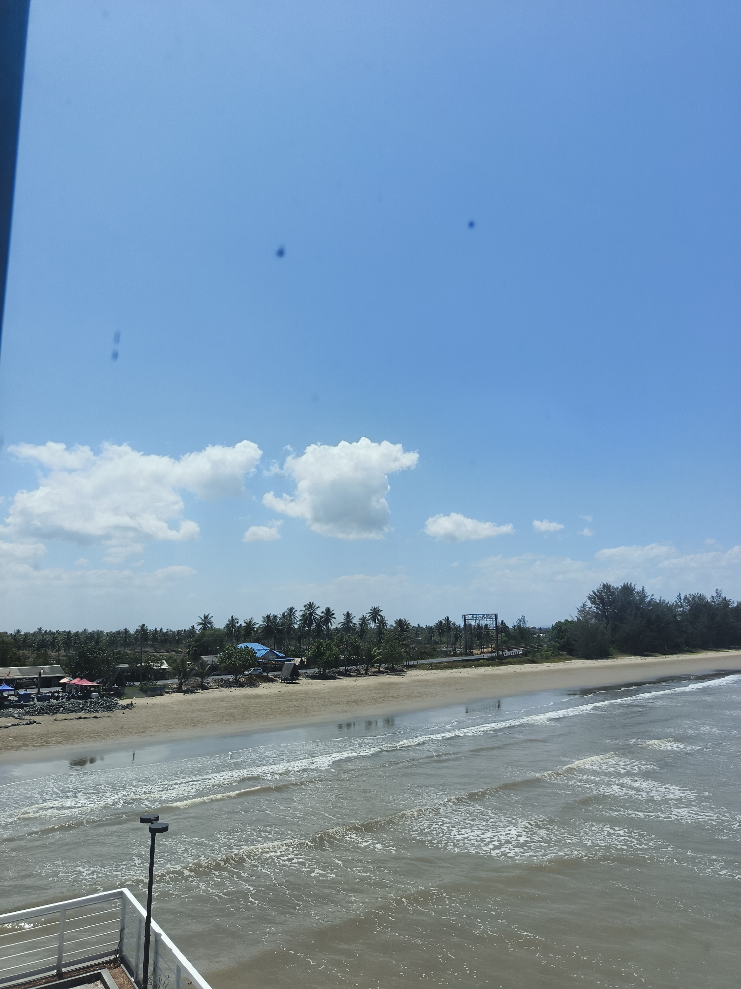 This is an outdoor, eye-level, wide shot of a beach and the ocean under a clear blue sky with some white clouds. The beach is sandy and lined with palm trees. There are a few small buildings and tents on the beach, suggesting a populated area. In the foreground, there is a white railing and a lamppost. The ocean has small waves crashing on the shore. The image appears to be taken from a slightly elevated position.