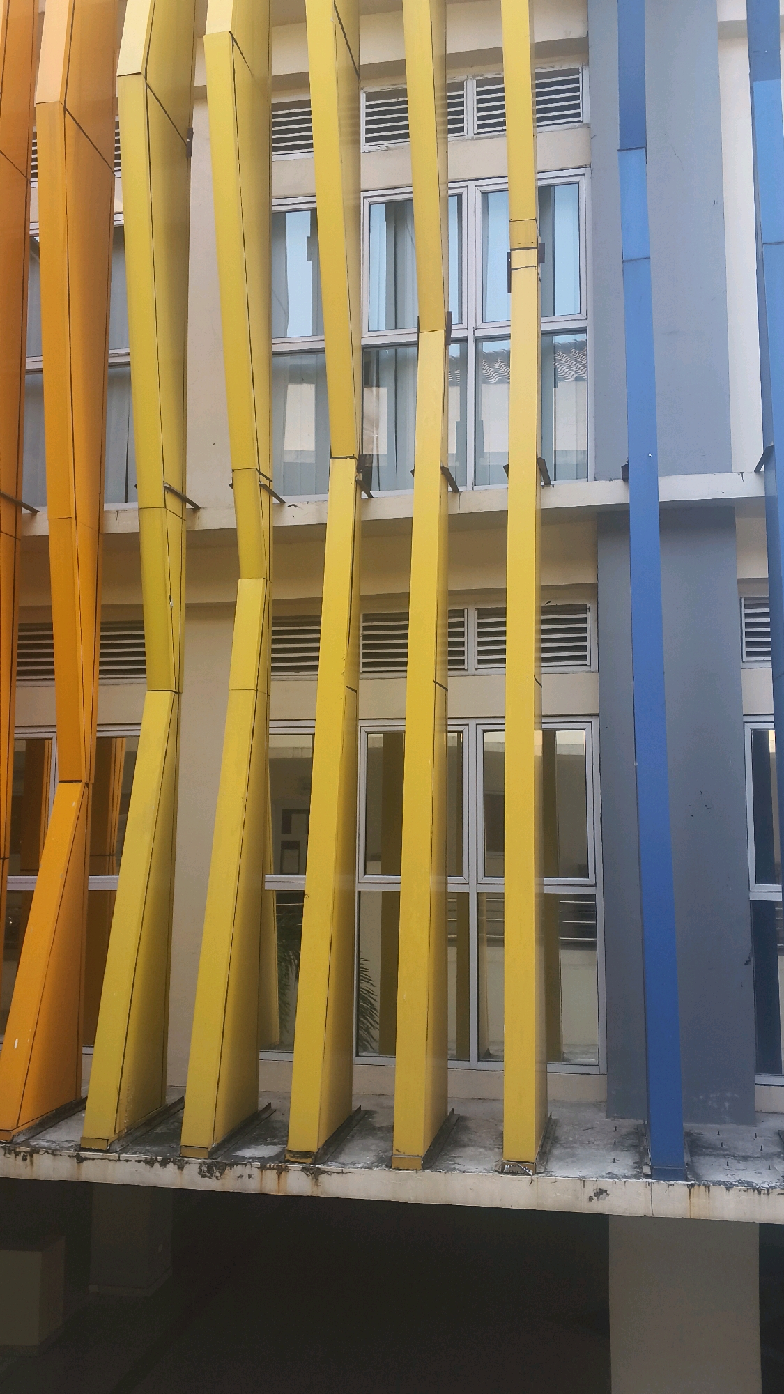 The image shows a brightly colored, modern building facade in Lohbener, Indonesia. The facade features a repeating pattern of vertical, angled panels in shades of orange, yellow, and blue, interspersed with windows. The yellow panels are prominent and appear to be made of metal, with a matte finish. They are angled inwards towards the building, creating a sense of depth. The windows are large and rectangular, with white frames, and reflect the sky. Some of the windows have horizontal louvers. The building appears to be multi-story, with a concrete walkway or ledge in the foreground. The overall impression is one of contemporary architecture and vibrant urban design.