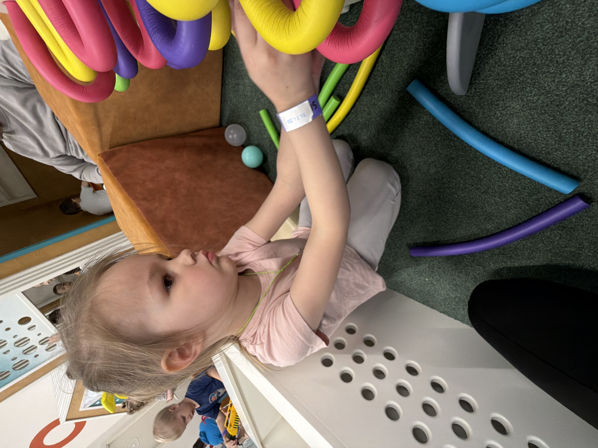 A young girl wearing a pink shirt and gray pants, and a bracelet, stands in an indoor play area. She has fair skin and blonde hair, and is reaching up toward colorful, flexible, tubular structures of pink, yellow, and purple. In the background, there are other adults and children. Two balls, one light gray and one light blue, rest on a brown surface. Blue, green, and purple tube segments are scattered on a green carpet. The environment includes furniture with circular cut-outs, which suggest the scene is taking place indoors at a play facility, possibly in a city such as Izhevsk, Russia, though that cannot be conclusively determined from the image.