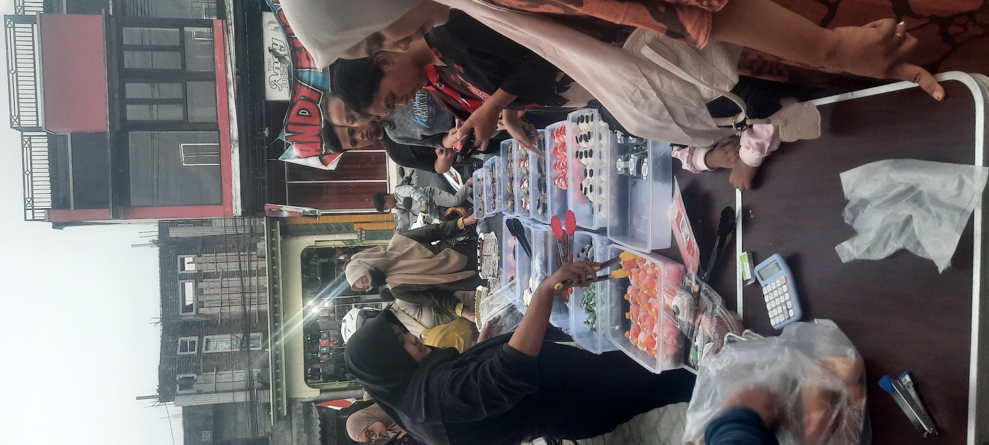 An outdoor market scene is depicted in Selopuro, Indonesia, with a focus on a table laden with various food items. Several individuals are engaged in browsing and purchasing goods. In the foreground, a woman in a black hijab is serving food from a clear plastic container using tongs. The container holds small, pink-colored food items topped with orange pieces. To her right, another woman in a beige hijab is looking at the food. Behind them, a man in a black t-shirt and a young boy on a scooter are visible. The table itself displays an assortment of pastries, cakes, and other desserts in clear plastic containers. Some of these items appear to be decorated with intricate frosting designs. A calculator, a stapler, and plastic bags are also present on the table. The background shows buildings, including one with red and grey sections, and flags. The overall atmosphere suggests a busy marketplace setting on an overcast day.
