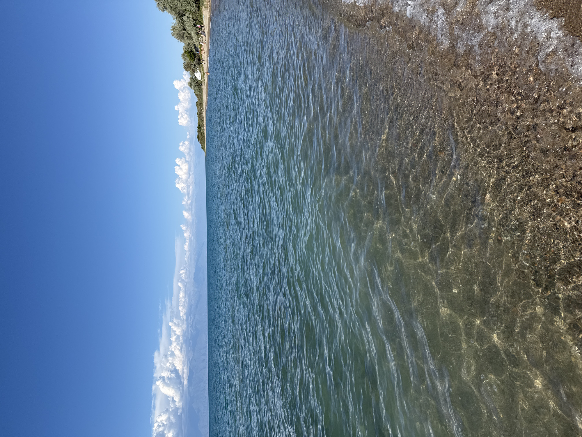 The image captures a serene view of a beach and the Issyk-Kul lake in the city of Balykchy, Kyrgyzstan. The foreground is dominated by the clear, shallow waters of the lake, revealing a pebbly bottom with sunlight shimmering through the ripples. The shoreline is a mix of sand and small stones, with gentle waves lapping at the edge.
In the middle ground, the vast expanse of the Issyk-Kul lake stretches towards the horizon, its turquoise waters reflecting the clear blue sky. A line of distant mountains, partially shrouded in clouds, forms a majestic backdrop. Along the shoreline to the right, a sandy beach is dotted with a few trees and some people are visible enjoying the day. There are a few scattered white tents, suggesting a camping or recreational area. The overall atmosphere is peaceful and idyllic, characteristic of a summer day at the lake.