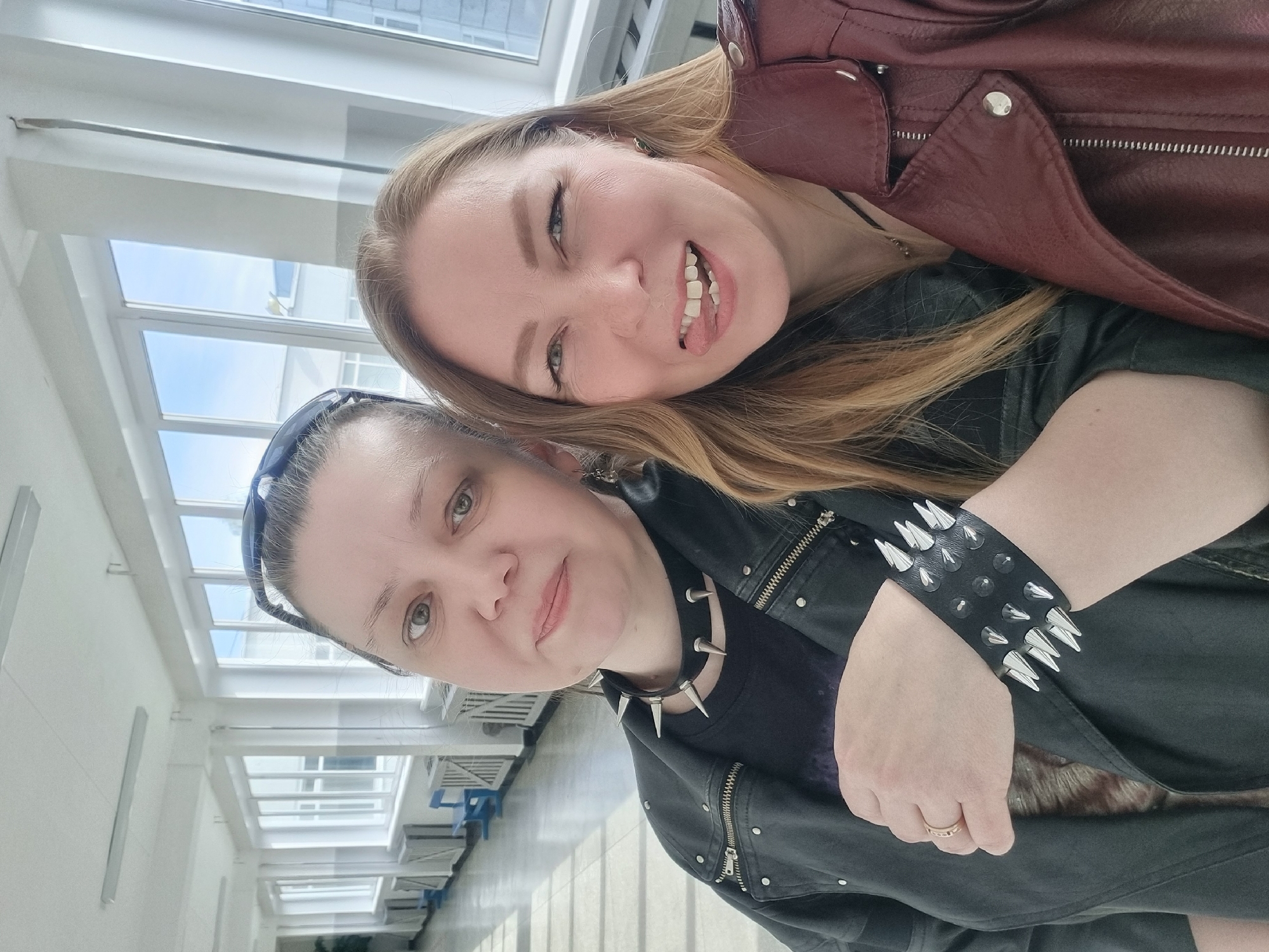 Two women are posing for a selfie indoors. The woman in the foreground wears a black leather jacket with zippers, a black spiked collar, and a studded wristband. The woman in the background has long blond hair and wears a red leather jacket. They are standing inside a building with large windows. Based on prior information, this location is the city of Chaikovsky, Russia.