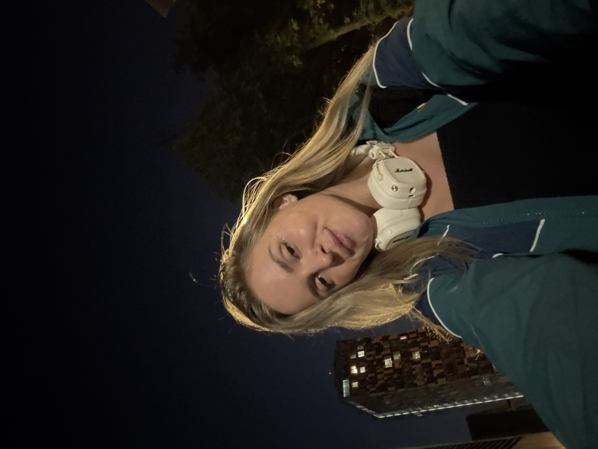 A woman is taking a selfie at night in Moscow, Russia. She has blonde hair and is wearing headphones around her neck. The background shows a tall building with illuminated windows and trees. The overall lighting suggests it is nighttime.