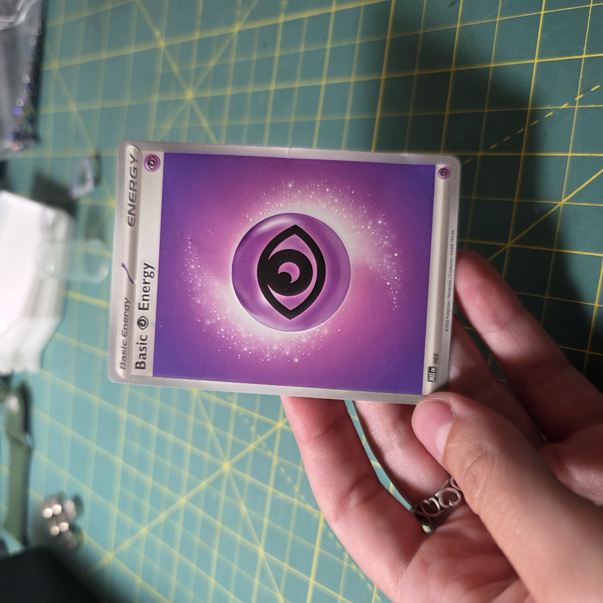 A person's hand, wearing a ring with a heart-shaped pattern, holds a "Basic Psychic Energy" Pokémon card. The card features a purple background with a central eye symbol. The card is positioned over a green cutting mat with yellow grid lines. In the background, out of focus, are several small metallic objects, a stack of white paper products, and a partially visible bag of small items. The scene appears to be indoors, possibly in a crafting or hobby area, and no specific location in Portland, United States of America can be determined from the visual information.