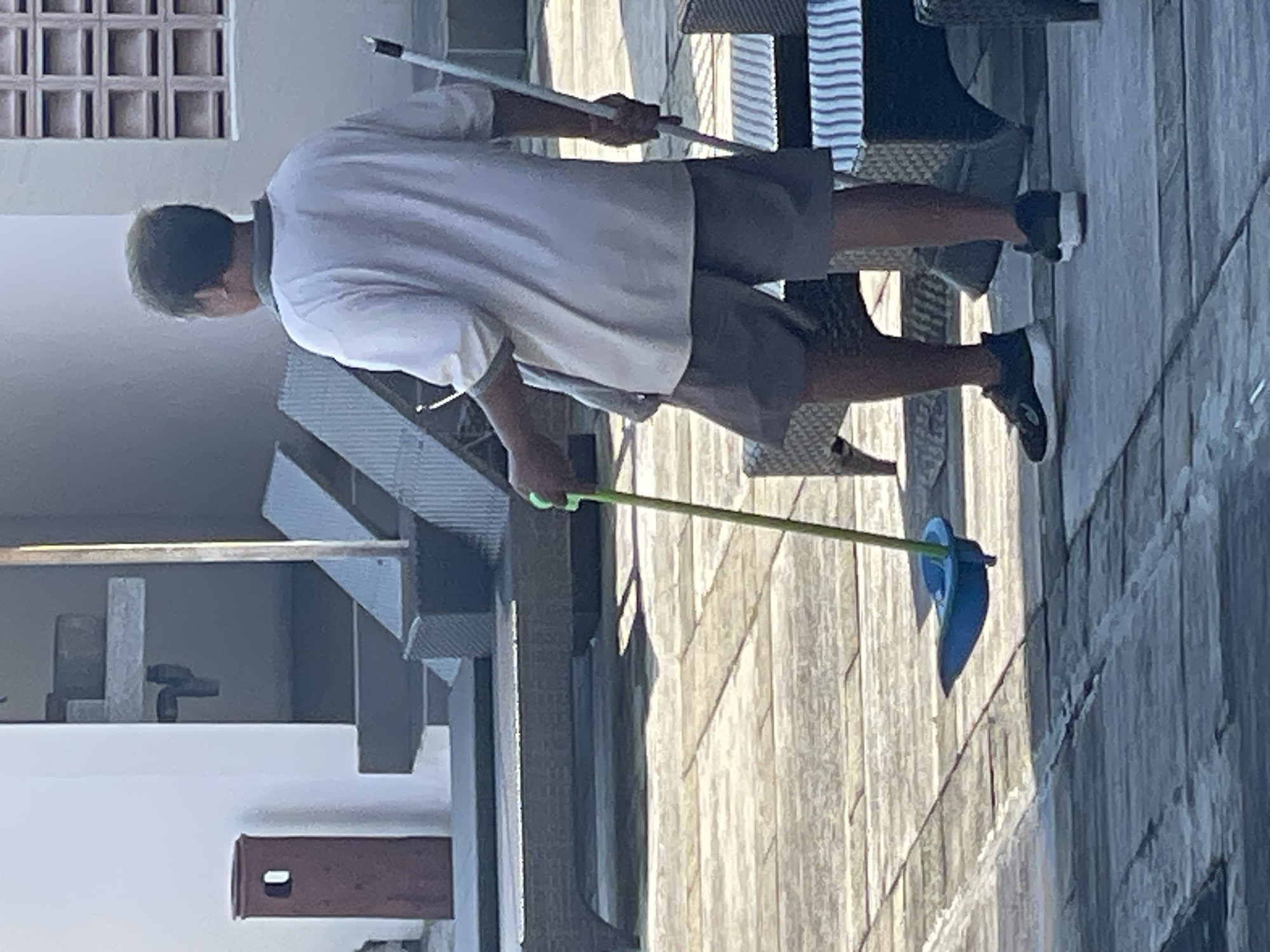 In Kuta Selatan, Indonesia, a man is standing on a tiled outdoor patio, holding a broom and a dustpan. He is wearing a light-colored polo shirt and dark shorts, with dark sneakers. The man appears to be cleaning the area. The background includes outdoor furniture, stairs, and a white wall with a window.