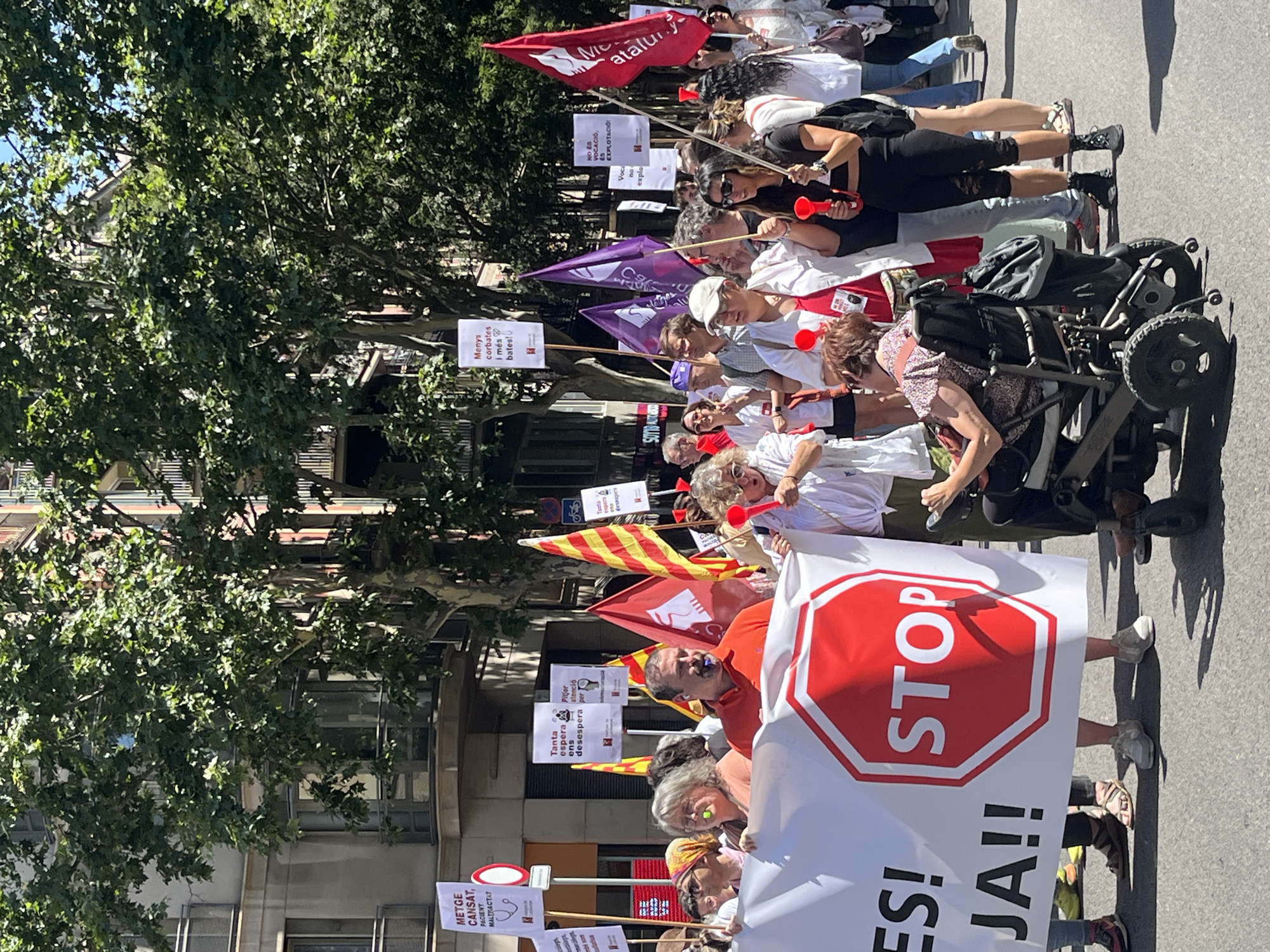 A group of protesters are marching down a street in Barcelona, Spain, on a sunny day. They are holding banners and flags, and some are wearing red items or blowing red trumpets. One banner in the foreground features a prominent red stop sign symbol with the word "STOP" printed inside. Another banner to the left of the stop sign has the words "ES!" and "JA!!" written on it. The protesters appear to be of various ages and backgrounds, and some are holding up signs with text in Catalan. In the background, there are buildings with balconies and trees lining the street. A woman in a wheelchair is participating in the protest. The overall atmosphere suggests a demonstration or rally.