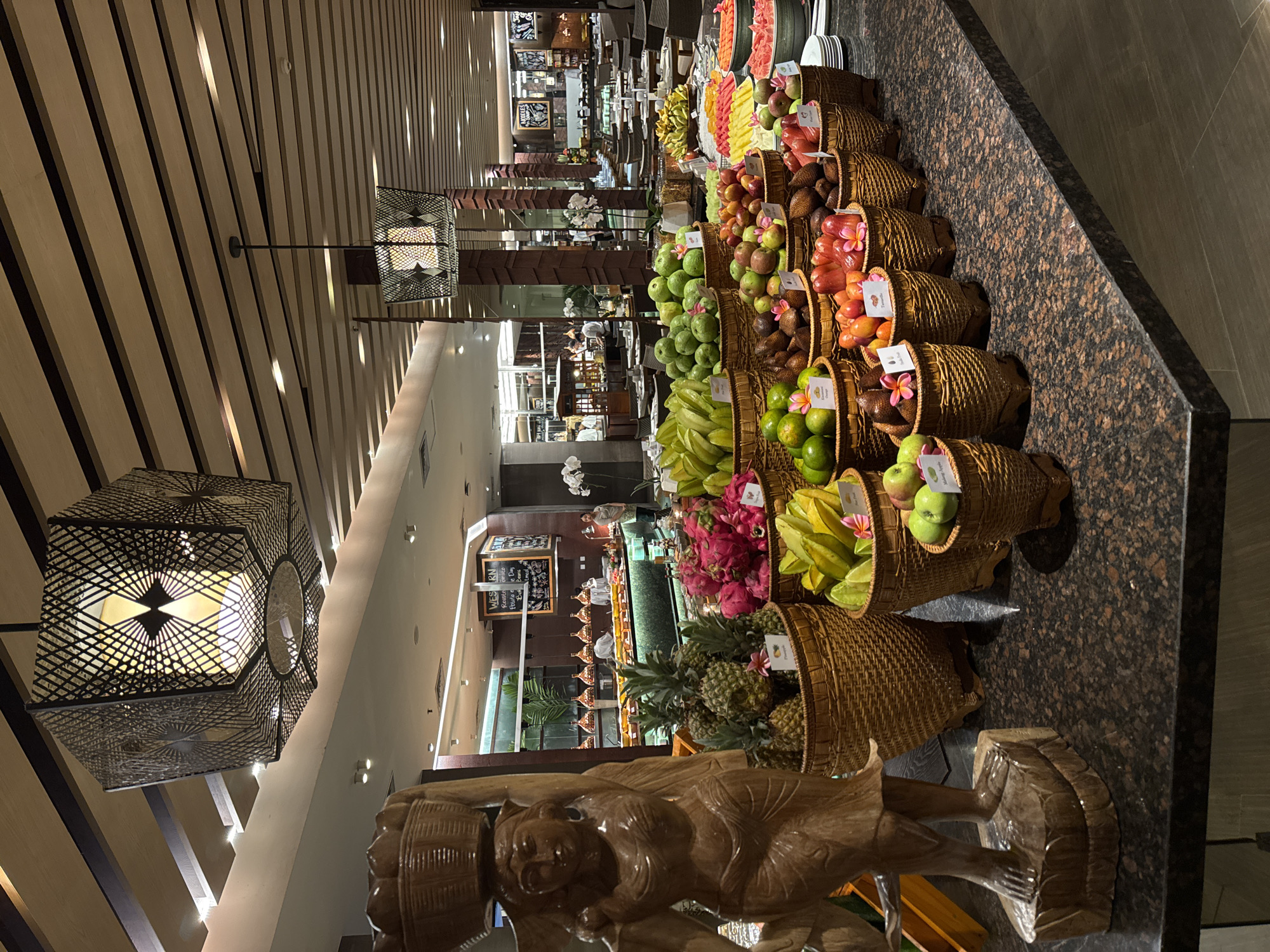 The image shows a buffet table laden with a wide variety of fresh fruits, presented in woven baskets. The fruits include pineapples, dragon fruits, starfruits, green apples, and what appear to be rambutan and mangosteen. In the foreground, to the left, is a carved wooden statue of a woman carrying a basket on her head. Behind the fruit display, a buffet line with food and serving stations is visible, with staff members attending to the setup. Several tables and chairs are set up in the dining area beyond the buffet. The lighting suggests an indoor setting, likely a restaurant or hotel dining area. The scene is located in Kuta Selatan, Indonesia, within a well-lit, spacious establishment.