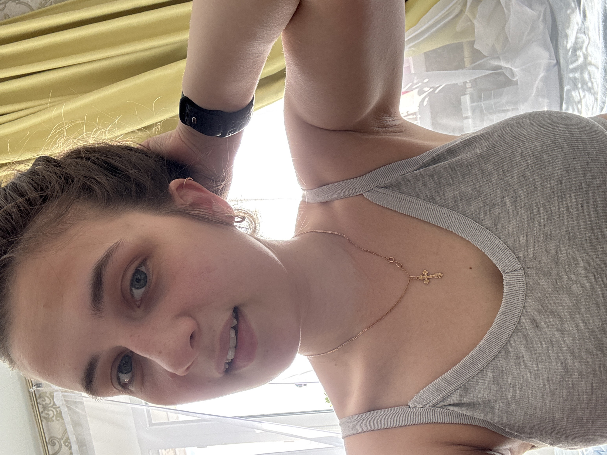A young woman is photographed indoors. She is wearing a gray tank top and a black smart watch on her left wrist. She is also wearing a thin gold necklace with a cross pendant. The background includes a yellow curtain and a window. The location is Chaikovsky, Russia.