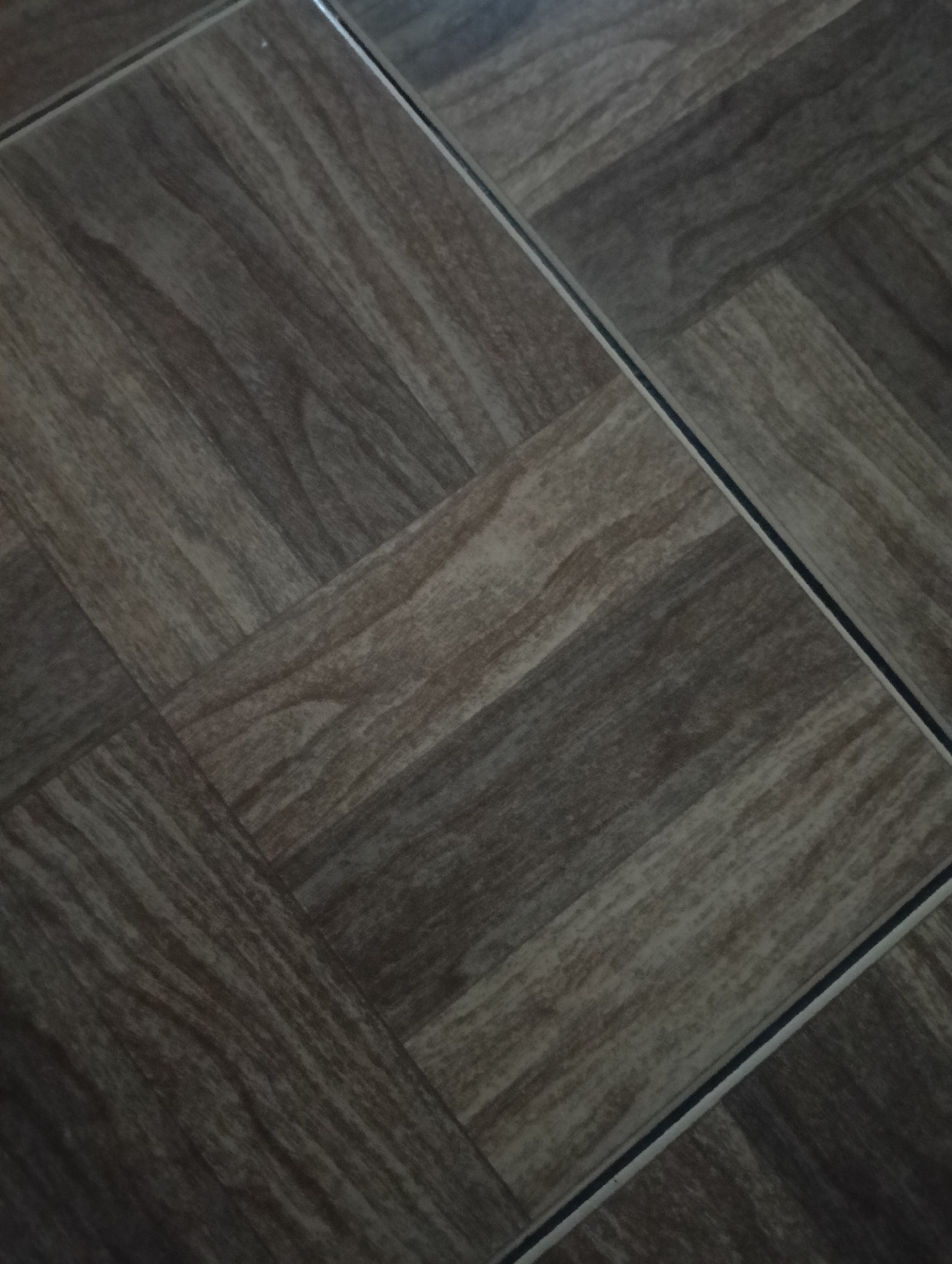 The image shows a close-up, overhead view of a tiled floor with a wood-grain pattern. The tiles are rectangular and arranged in a herringbone or basketweave pattern, with dark grout lines separating them. The color of the tiles is a mixture of dark brown and gray, creating a natural wood appearance. The lighting suggests an indoor setting, possibly a residential or commercial space in Adinuso, Indonesia. There are no people or specific objects visible in the frame, only the texture and pattern of the flooring. The perspective is at an angle, with the floor tiles receding into the background.