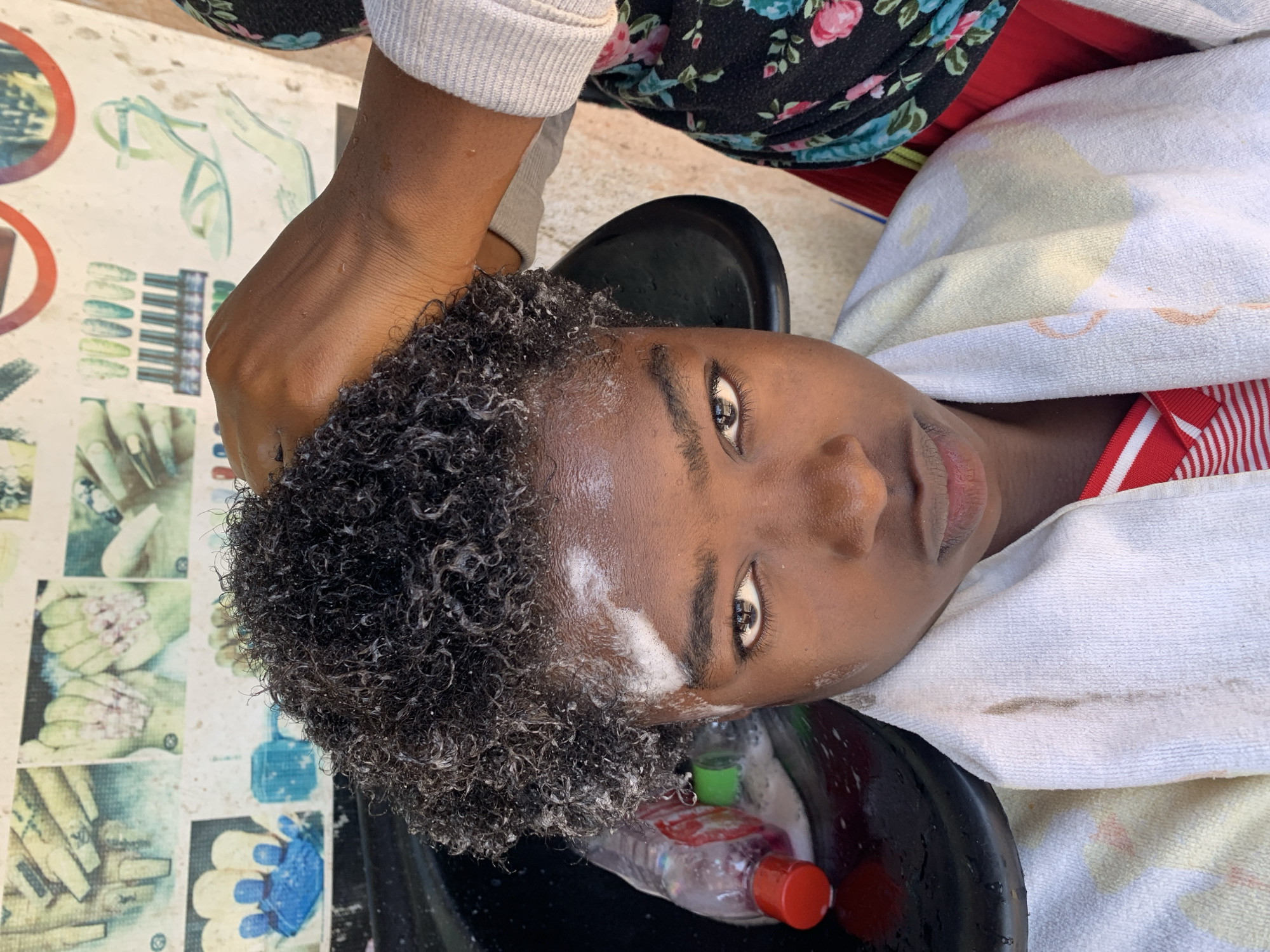 In Rafin Par, Nigeria, a close-up shot focuses on a young person receiving a hair wash in a black basin. Their curly, dark hair is wet and appears to be undergoing some form of treatment, with white foam visible on their forehead and around their hairline. A hand, belonging to someone off-camera, is seen gently working through the hair. A partially obscured plastic bottle, likely containing hair product, is visible in the basin to the left. The person is wearing a white towel draped around their shoulders, and their red and white striped shirt is partially visible. Behind them, a wall displays a collage of images, including pictures of fingernails, nail polish bottles, and high-heeled shoes. The lighting suggests an outdoor or well-lit indoor environment.