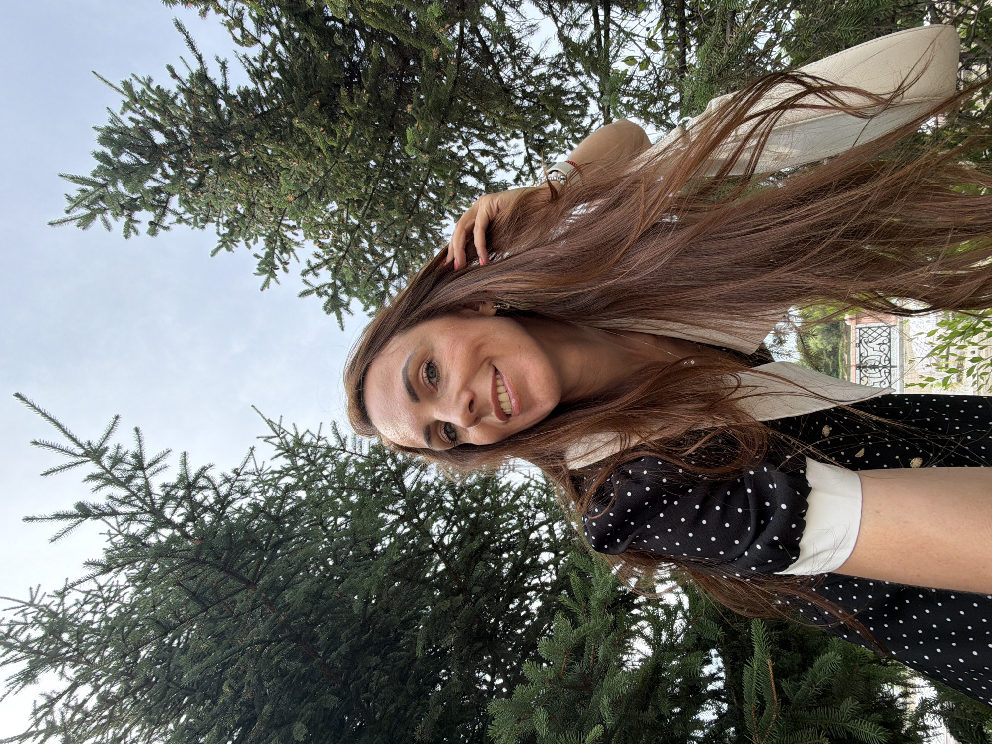 A medium shot captures a woman with long, auburn hair smiling as she touches her hair, positioned outdoors. She is wearing a black dress with white polka dots and a cream-colored shawl or cardigan. Her hair cascades down, obscuring much of her body. In the background, lush green fir trees dominate the frame, with a pale, overcast sky visible above. The composition is framed from a low angle, looking up at the woman and the trees. The location appears to be a park or garden setting within Birobidzhan, Russia.