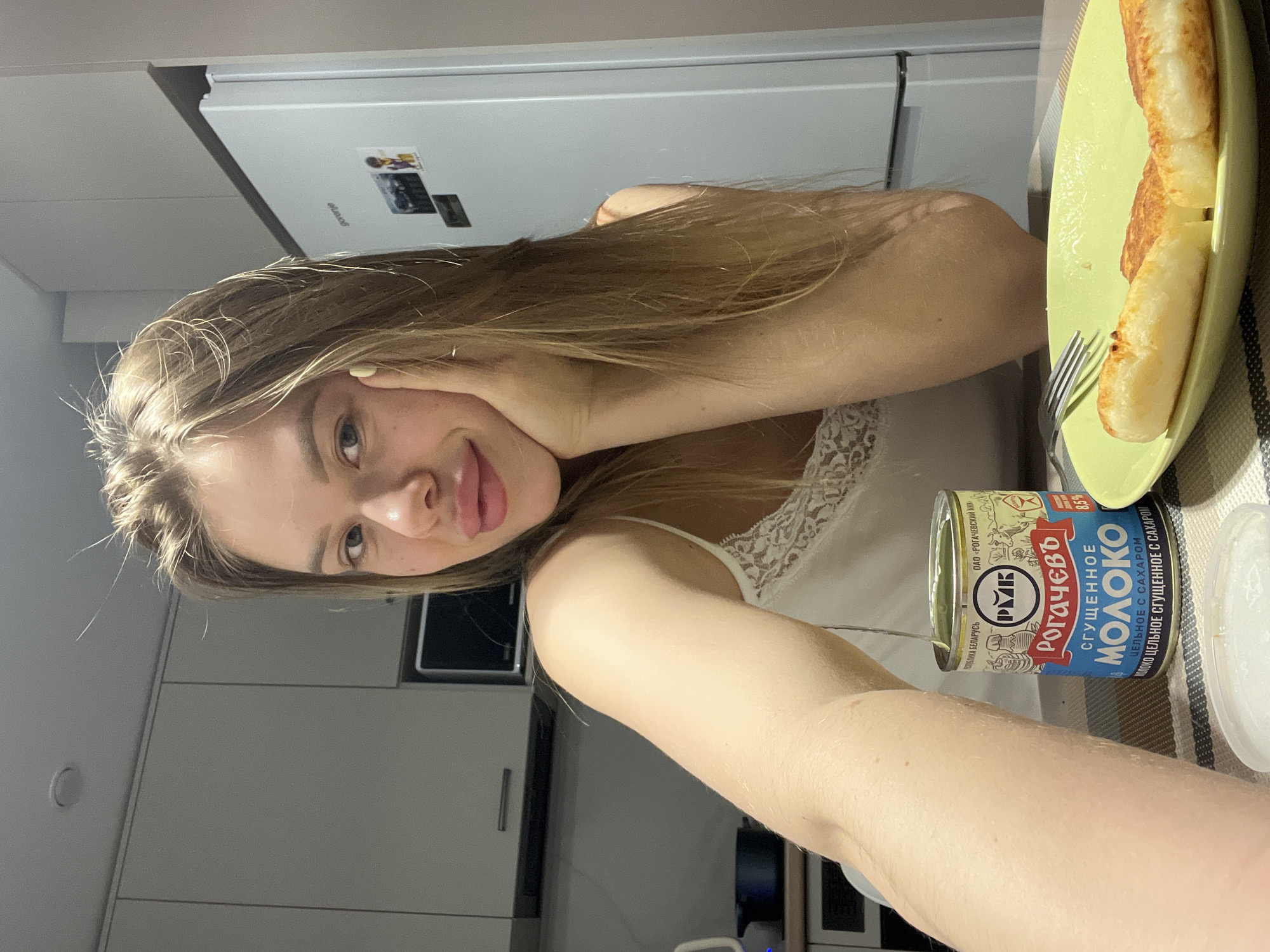 A young woman with long blonde hair is facing the camera, holding up a can of "Rogachev" condensed milk. She is in a kitchen with white cabinets and appliances. On the counter, there is a green plate with what appears to be fried food and a fork. The location is the city of Chaikovsky, Russia.