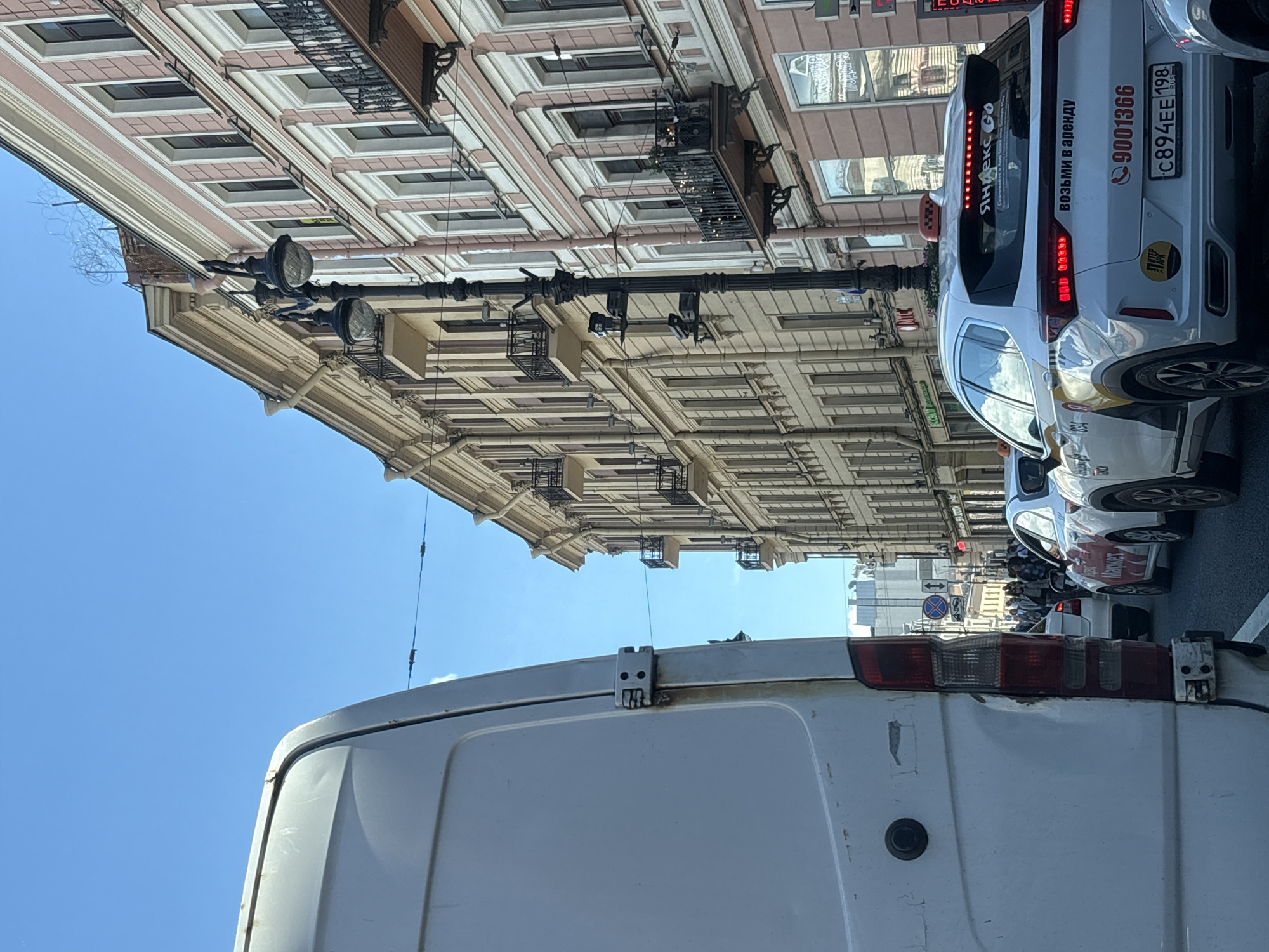 The image depicts a traffic scene on a street in Saint Petersburg, Russia. The perspective is from inside a white van, looking forward and slightly to the right. The street is lined with multi-story buildings with classical architecture, featuring balconies and ornate detailing. The sky is clear and blue. In the foreground, the rear of the white van is visible. Several cars are in traffic ahead, including white sedans with taxi markings. One taxi has "Яндекс GO" (Yandex Go) written on its rear window and "Возьми в аренду" (Take for rent) written on its trunk. The license plate of this taxi is C894EE 198 RUS. A lamppost with multiple lights stands on the sidewalk between the buildings. Pedestrians are visible on the sidewalk and crossing the street in the distance. The overall impression is of a typical urban street scene during daylight hours.