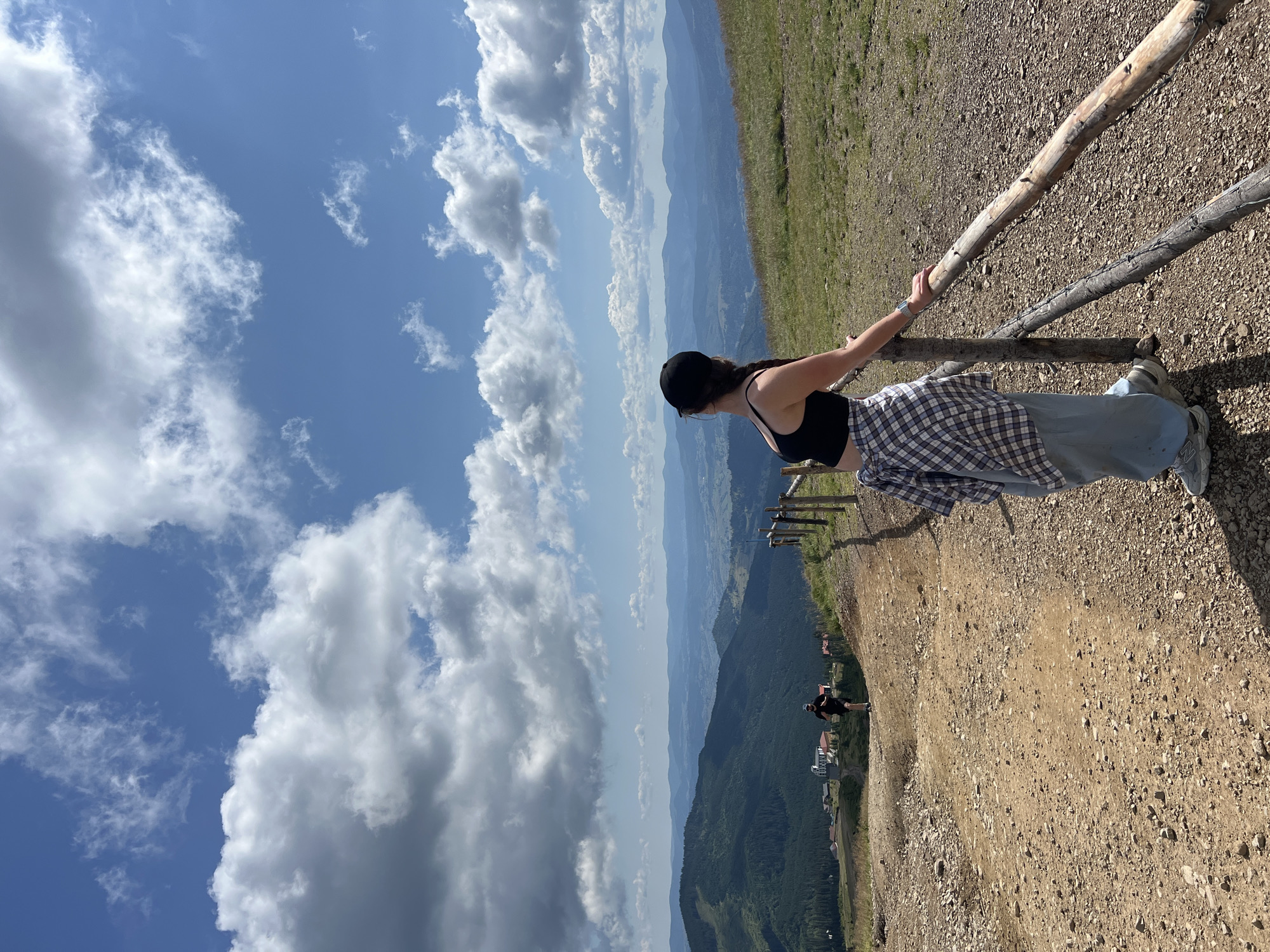 In the city of Chorna Tysa, Ukraine, a young woman stands on a dirt path, leaning on a rustic wooden fence. She wears a black crop top, light blue pants, a plaid shirt tied around her waist, and a black baseball cap. Her hair is in a braid that hangs over her shoulder. She looks out at a breathtaking landscape of rolling green hills and distant mountains under a bright blue sky dotted with fluffy white clouds. Further down the path, another person is visible, appearing to be running. The environment suggests a mountainous or hilly terrain, possibly a hiking trail or a scenic overlook. The ground is covered in dirt and small stones, with patches of dry grass on the slopes. The wooden fence is weathered and appears to be made of rough-hewn logs and branches. The scene is captured in daylight, with clear visibility.