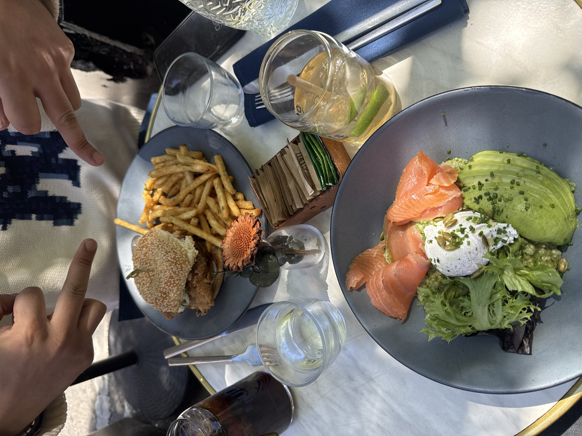 This bright daylight, high-angle photograph captures a casual meal shared by at least two individuals at what appears to be an outdoor cafe or restaurant. The light-colored table surface, dappled with sunlight, is laden with food, drinks, and tableware.
Two sets of hands are visible. One hand, with a light skin tone, points an index finger towards the left plate, seemingly drawing attention to the dish. Another hand, also with a light skin tone and wearing a light-colored bracelet, has its middle finger extended upwards towards the lower left portion of the frame. This gesture, within the context of a shared meal, likely suggests a playful or informal interaction between dining companions.
On the left, a dark blue/grey plate holds a burger, likely fried chicken, served on a sesame seed bun, accompanied by a generous portion of golden french fries. A small, dried orange flower is placed decoratively on this plate. On the right, a similar dark blue/grey plate features a healthy brunch item: several slices of smoked salmon, fanned slices of avocado, a poached egg topped with chives and pumpkin seeds, and a base of mixed green lettuce.
Beverages include a clear glass with a light-colored drink, ice, and lime wedges, and another clear glass containing a dark beverage, possibly iced tea or soda. An empty clear glass is also on the table. A small wooden caddy holds various sugar and sweetener packets, with some brown ones clearly labeled "RAW." Forks are visible on a blue napkin, and a small vase with a dried flower and leaves sits between the plates. The scene suggests a relaxed dining experience during daytime, consistent with a brunch or lunch outing in a location like New York, United States of America.