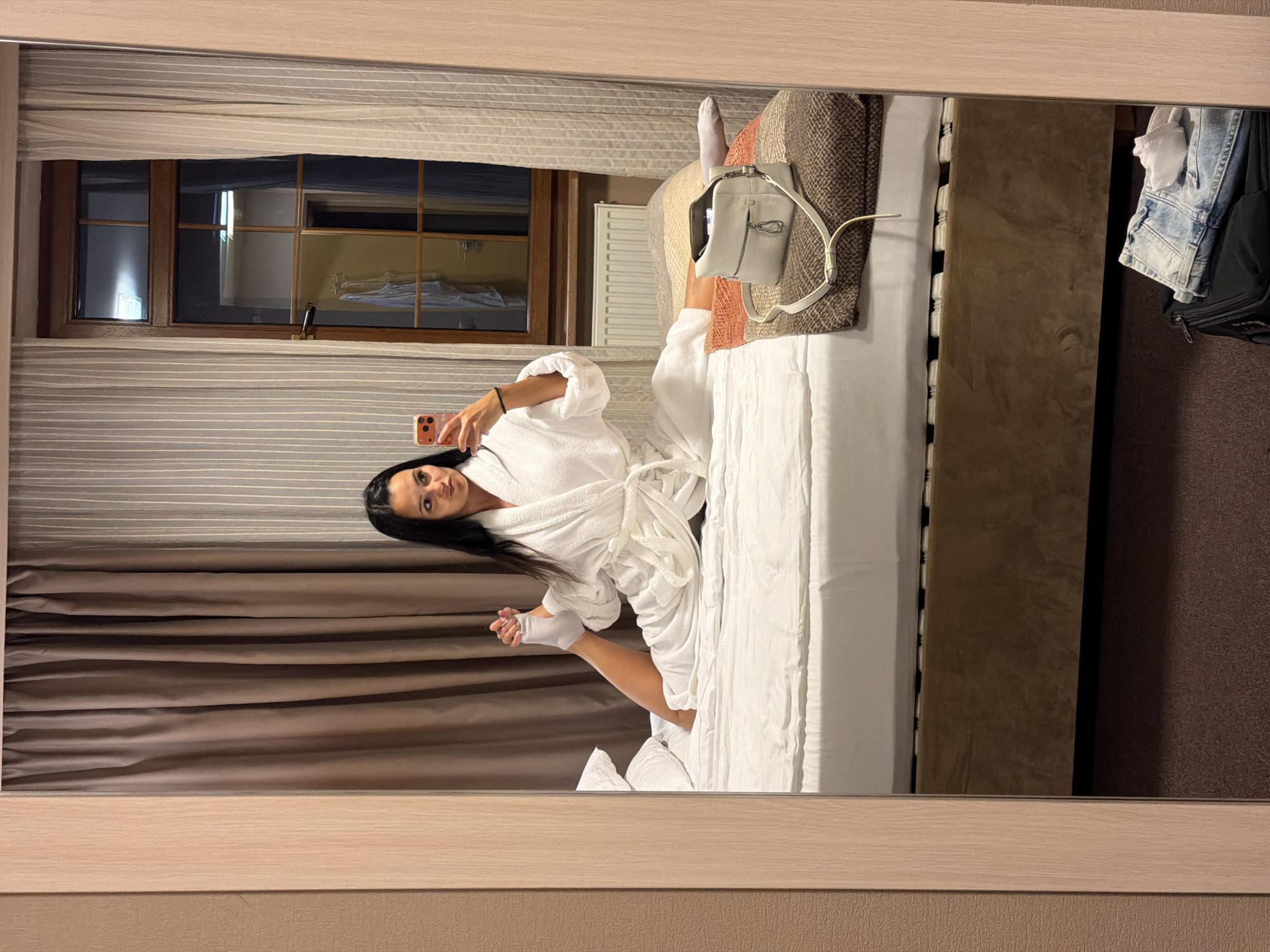 A young woman with long dark hair is performing a split on a hotel bed, wearing a white bathrobe and white socks. She is taking a selfie with her smartphone. A white handbag is placed on a folded blanket next to her. A suitcase with folded jeans and a white cloth is on the floor in the foreground, visible in the reflection of a mirror. The room has curtains, a window with a view of another room, and a radiator. The location is a hotel room in Svetlogorsk, Russia.