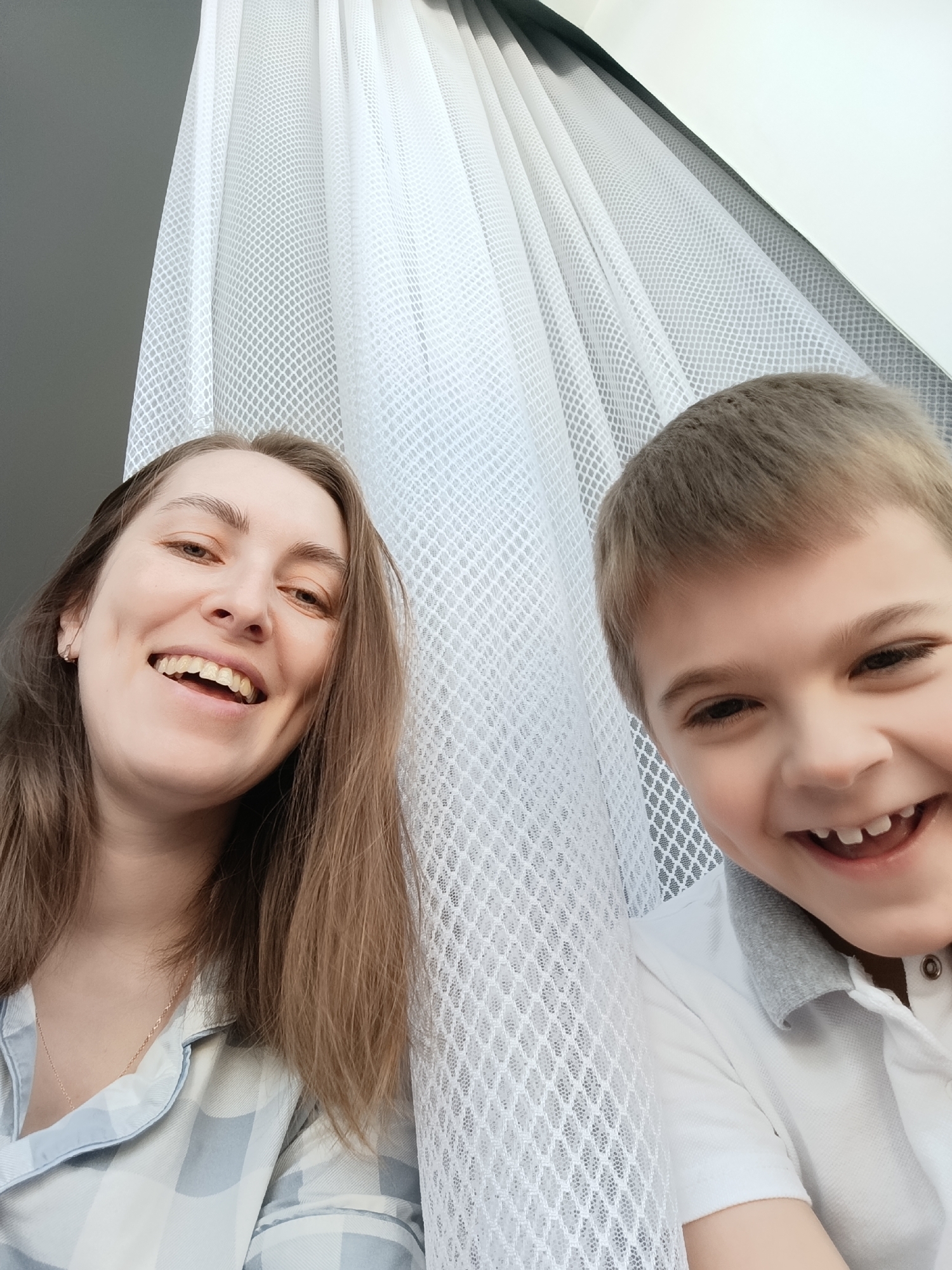 The image shows a selfie of a woman and a boy. The woman is on the left, wearing a light blue and white checkered shirt, and has long brown hair. She is smiling broadly, revealing her teeth. The boy is on the right, wearing a white polo shirt with a gray collar. He is also smiling broadly, showing his teeth. Behind them is a white, net-like curtain. The lighting appears to be natural and bright. Given the user's request for specificity, and the lack of visual cues, it is impossible to confirm or deny the location in Chaikovsky, Russia based solely on the provided image. There are no additional items or actions visible that contribute to a precise location or context.