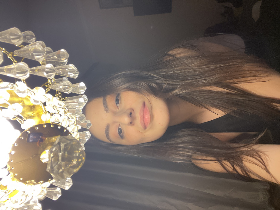 An indoor scene features a young adult female positioned beneath a brightly illuminated chandelier. The specific city location, Moscow, Russia, cannot be determined from the visual evidence.
The female subject, centered in the lower two-thirds of the frame, possesses long, straight, dark brown hair, brown eyes, and light-to-medium skin tone. Her lips are a natural pink, with a slight upturn at the corners, and she looks directly into the camera. She wears a dark-colored, sleeveless top.
Dominating the upper portion of the image is a multi-tiered chandelier constructed from brass-toned metal and adorned with numerous clear, teardrop-shaped glass or crystal pendants. The chandelier is illuminated, acting as a primary light source. A reflection consistent with a camera lens is visible on a metallic component of the chandelier in the upper-left area.
The background is dimly lit. To the left, dark, vertically textured fabric, likely curtains, is visible. To the right, the environment is indistinct and dark, suggesting an indoor room setting. The lighting originates from the overhead chandelier, casting direct light on the subject and the fixture itself, while the background remains in shadow.