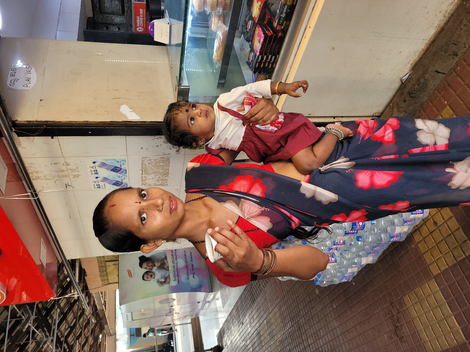 A woman holding a baby stands on a street in an urban area of Unknown, Unknown, next to a building. The woman has dark hair pulled back, brown skin, and a bindi on her forehead. She is wearing a dark sari with a red floral pattern, gold bangles, and a gold nose ring. She holds a white cup in her left hand. The baby has dark hair and brown skin. The baby is wearing a white shirt and a red dress, with gold bangles on its left wrist. The baby's left leg has a silver anklet. To the right of the woman are stacks of bottled water. Behind the woman is a building that appears to be a restaurant or bakery. A window sign reads "PURE VEG." There is a sign for Madras Coffee House. A sign for baby care products is partially visible, as well as advertising for cookies (Treat). The street has brown and gold brick-like flooring.