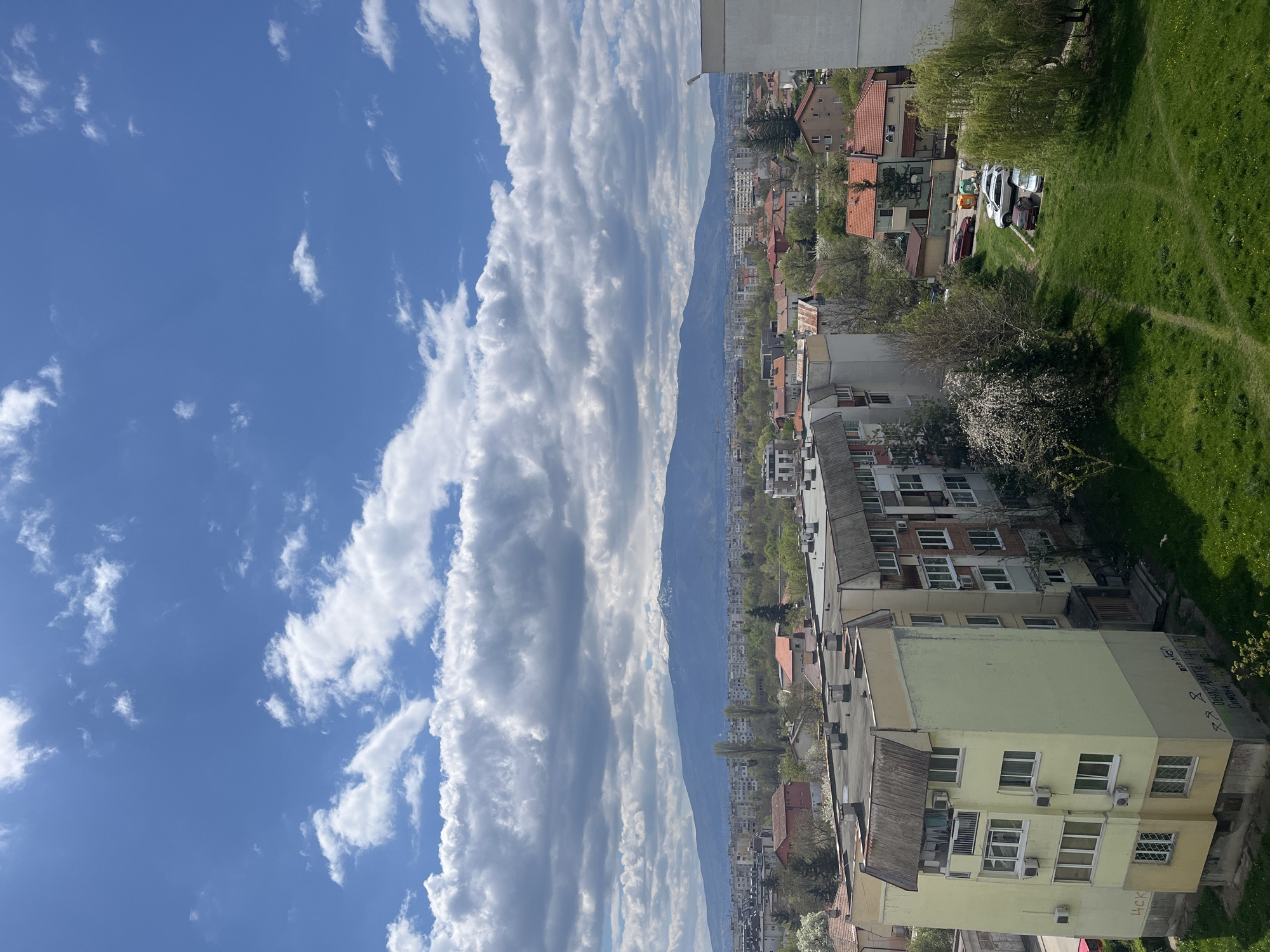 The image displays a panoramic view of a city, identified as Sofia, Bulgaria, set against the backdrop of a distant mountain range, likely Vitosha Mountain. The perspective is from an elevated vantage point, showcasing a sprawling residential area under a vibrant blue sky.
The sky is a prominent feature, adorned with large, white cumulus clouds arranged in distinct, elongated bands stretching horizontally. The clear blue sky and bright sunlight suggest it is daytime, likely late morning or early afternoon, with fair weather conditions.
In the foreground and mid-ground, numerous multi-story residential buildings of varying architectural styles are visible. Many feature red-tiled roofs, while others have flat, gray ones. A distinctive yellow/light green building stands out in the mid-right section. Its facade is marked with graffiti, including Cyrillic text that appears to read "ОБЕДИНЕНИ ЦСКА" (United CSKA), accompanied by stars and an anarchy symbol. Several air conditioning units are mounted on the exterior walls of these buildings.
Interspersed throughout the urban environment are patches of lush green grass and various trees, some of which are in bloom with white blossoms, indicating a spring season. On the right side, a grassy area shows discernible tire tracks or well-worn paths, and a few cars are parked on an unpaved section near some garbage bins. The distant mountain range appears hazy blue, defining the horizon line. No specific activities or individuals are depicted; the image captures a tranquil, everyday urban landscape.