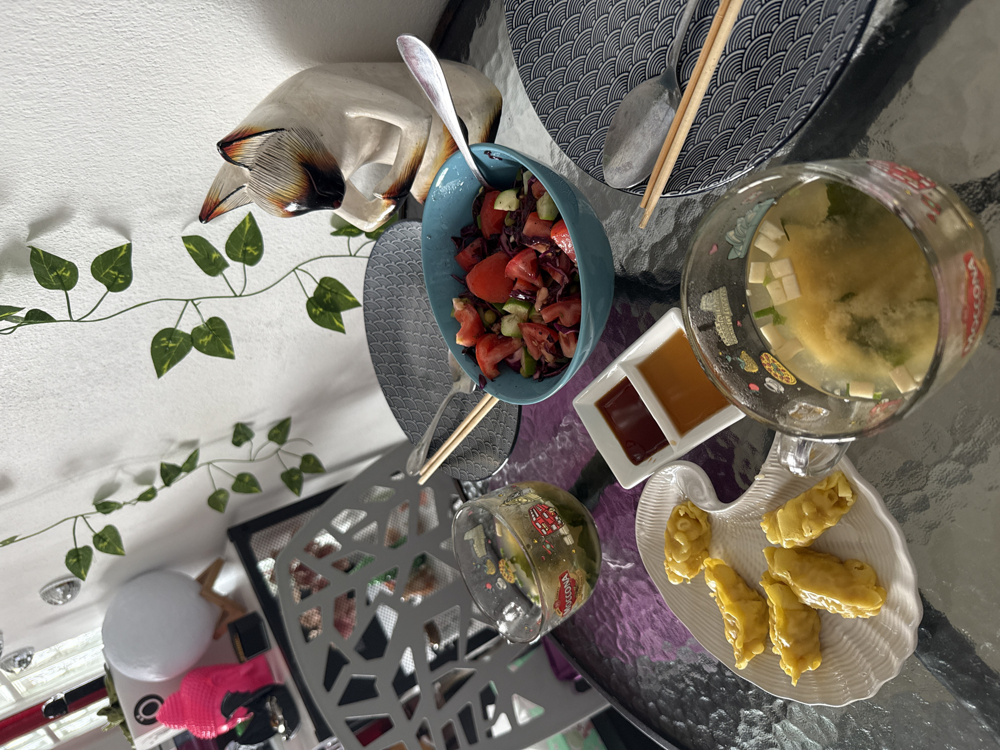 The image shows a table set for a meal, likely outdoors or in a well-lit room with natural light. The table is made of glass with a textured surface, possibly reflecting the purple placemat underneath. On the table are several food items and serving dishes. In a blue bowl, there is a salad consisting of chopped tomatoes, red cabbage, and cucumber. Adjacent to the salad are two small white dishes containing sauces, one appears to be red and the other amber.
A white, heart-shaped plate holds several golden-brown dumplings. Two clear glasses are present. One glass contains a yellowish liquid with green herbs and cubed white pieces, possibly a soup. The other glass, with graphics of London landmarks, also contains a similar liquid with green elements and white cubes.
There are two patterned plates with a blue and white wave design. One plate has chopsticks and a spoon resting on it, while the other plate is empty. A wooden cat figurine, with a Siamese cat-like appearance, is positioned behind the salad bowl, looking down. In the background, a white wall is decorated with artificial green vines. To the left, a grey chair with a geometric pattern is visible, and further left, a white lamp with wooden legs and a pink Buddha-shaped object are present.
This scene does not provide specific information to determine its location in the city of Ko Samui District, Thailand. The presence of the London-themed glass is a personal decorative choice and does not indicate the location.