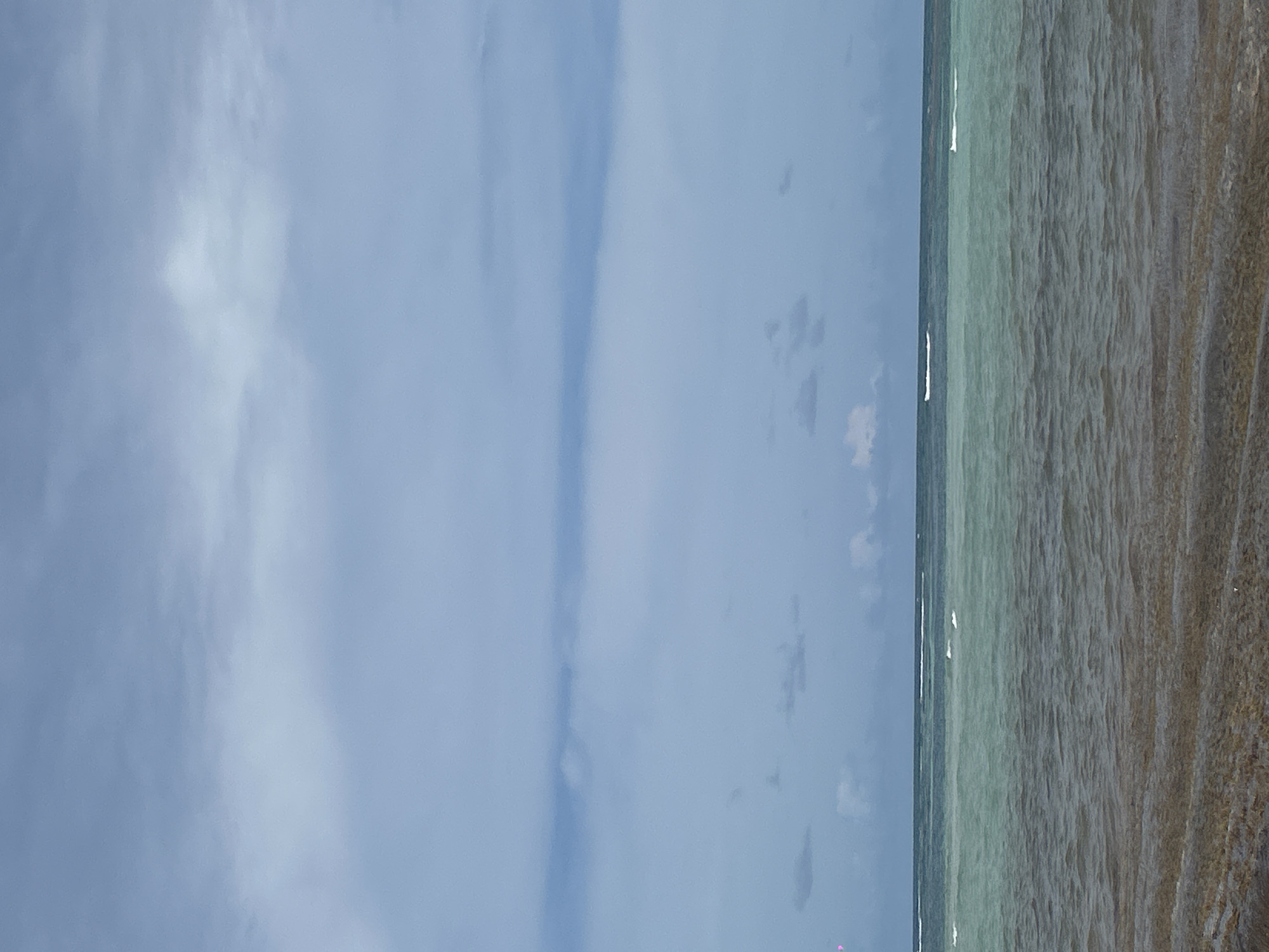 The image depicts a coastal scene in Sainte-Anne, France. The left side of the image shows a cloudy sky. The right side features the ocean meeting the shoreline. The water appears clear with a slightly greenish tint, transitioning to a sandy beach. Small waves are visible along the shoreline. No people or specific objects are clearly discernible in the scene.