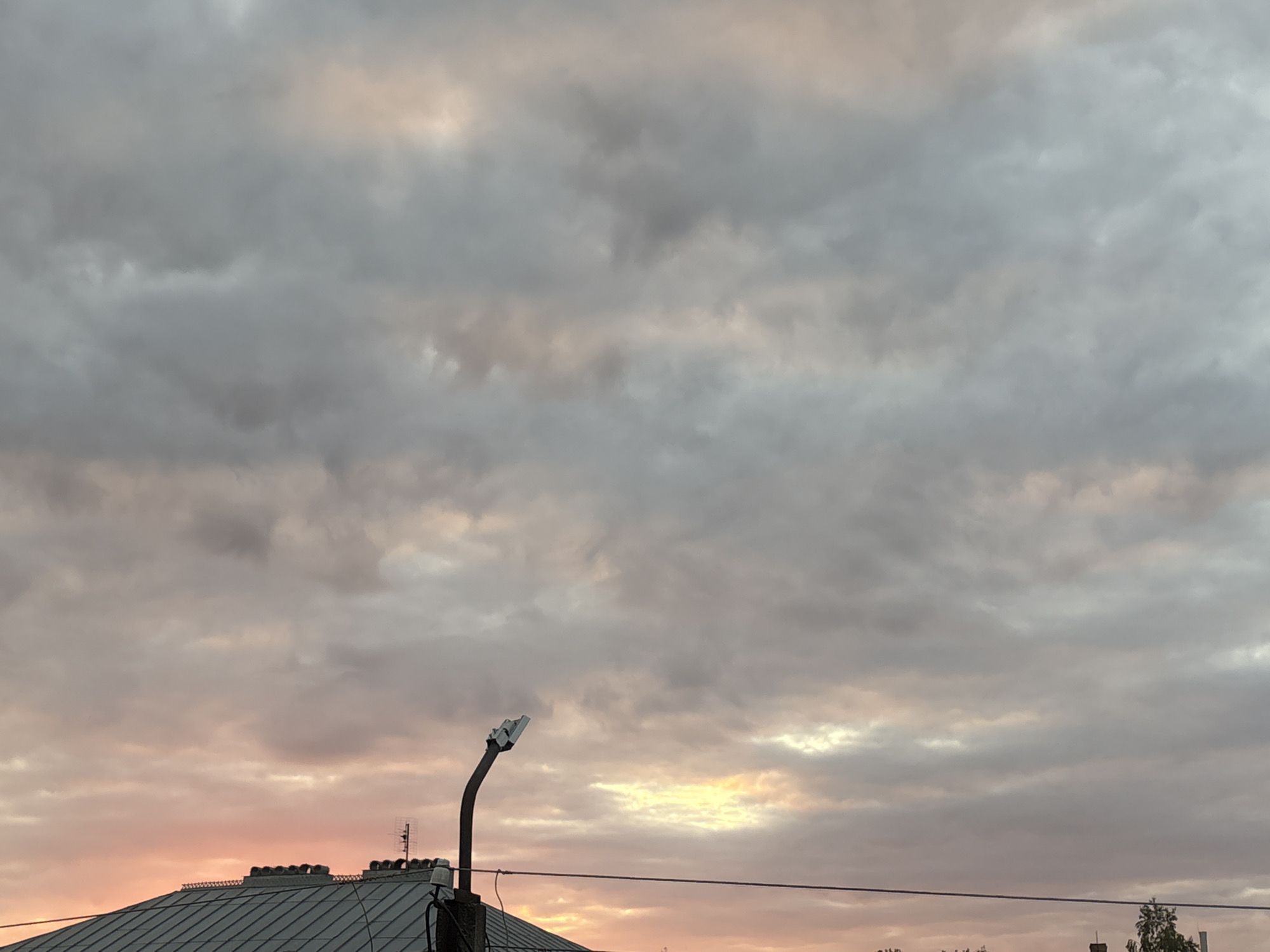 This image depicts a dusky sky over the city of Georgiyevsk, Russia. The sky is filled with a mixture of gray and pinkish clouds, suggesting either sunset or sunrise. In the foreground, the sloped, gray metal roof of a building is visible, topped with several chimney-like structures. A dark, curved lamppost with a light fixture extends upward from the roof. A thin wire runs horizontally across the frame, partially obscuring the view of the sky and buildings. The overall scene is overcast, with pockets of softer light breaking through the clouds.