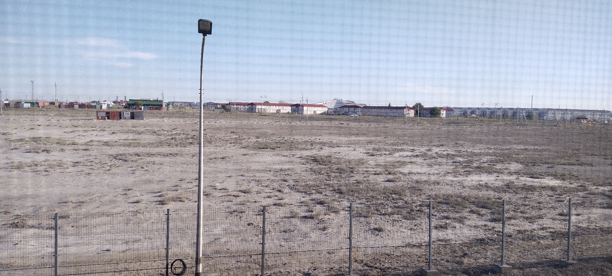 The image depicts an outdoor scene under a clear, pale blue sky, suggesting daytime with fair weather. The foreground features a chain-link fence, with metal posts and a coiled cable visible near the bottom right. Beyond the fence lies a large, sparsely vegetated, arid expanse of dry earth and scrubby plants.
In the middle ground, a tall, slender lamppost stands prominently on the left side, topped with a rectangular light fixture. Further in the background, a row of shipping containers, some colored red, green, and grey, are neatly arranged. Beyond the containers, a long, low-rise building complex with red roofs stretches across the horizon. The architecture of these buildings suggests a residential or communal living facility. In the far distance, more industrial or infrastructural elements, including what appear to be power lines or communication towers, are visible.
The overall setting appears to be an undeveloped or industrial area on the outskirts of a settlement, possibly a port or logistics hub, given the presence of shipping containers. The arid landscape and sparse vegetation are indicative of a dry climate. No people are visible in the image, and there is no discernible text. The scene conveys a sense of an industrial or administrative zone within a somewhat desolate landscape.