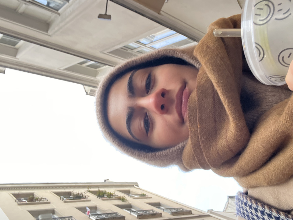 A low-angle selfie features a young female subject centrally positioned against an urban backdrop, specifically in Paris, France. She wears a light brown hooded garment and a darker brown scarf, looking directly at the camera with a slight smile. Her dark hair is visible beneath the hood, complemented by dark eyebrows and eyes. In the lower right foreground, her hand holds a translucent plastic cup with a lid and straw, decorated with black outline smiley faces and containing a light-colored beverage. The background consists of a narrow street flanked by two tall, light-colored buildings under an overcast sky. The building on the left exhibits several windows with ornate dark iron balconies, some adorned with small plants, and a red and white flag, indicative of Poland, displayed on an upper-level balcony. The building on the right shows similar architectural features, including windows and a wall-mounted light fixture. The overall architecture is characteristic of Parisian Haussmannian buildings.