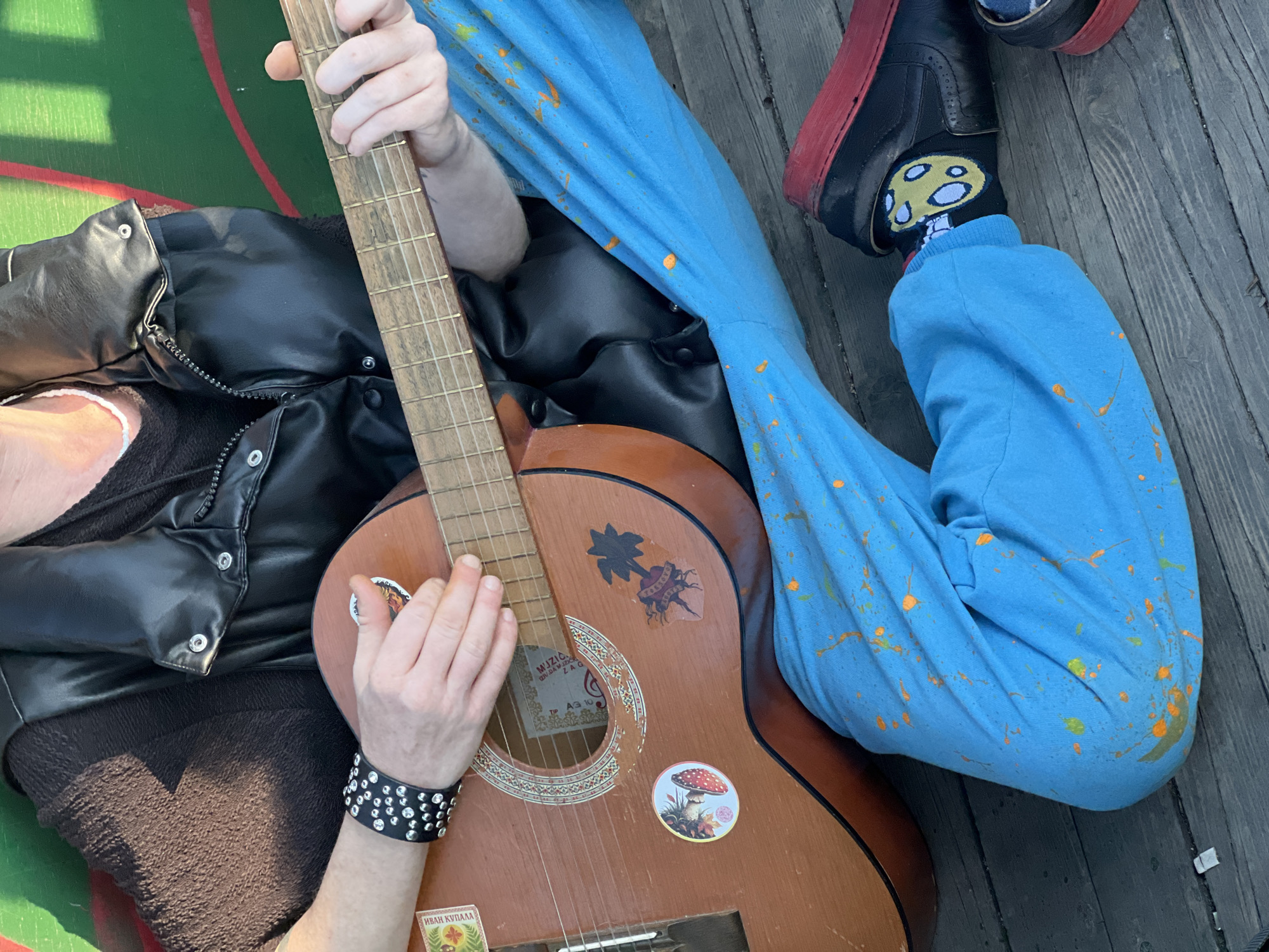 The image captures an individual seated, holding a guitar. The person is wearing blue pants with orange splatters, black socks with mushroom graphics, and black platform shoes with red soles. A black studded wristband adorns their left wrist. They are also wearing a black leather jacket. The guitar is brown and decorated with several stickers, including one with a palm tree and another with a mushroom. The individual is seated on what appears to be a wooden surface, possibly decking. Part of a green surface with red and white markings is visible in the background.
Given the information, the location is indeterminate; there are no visual clues that suggest this is Belgrade, Serbia.