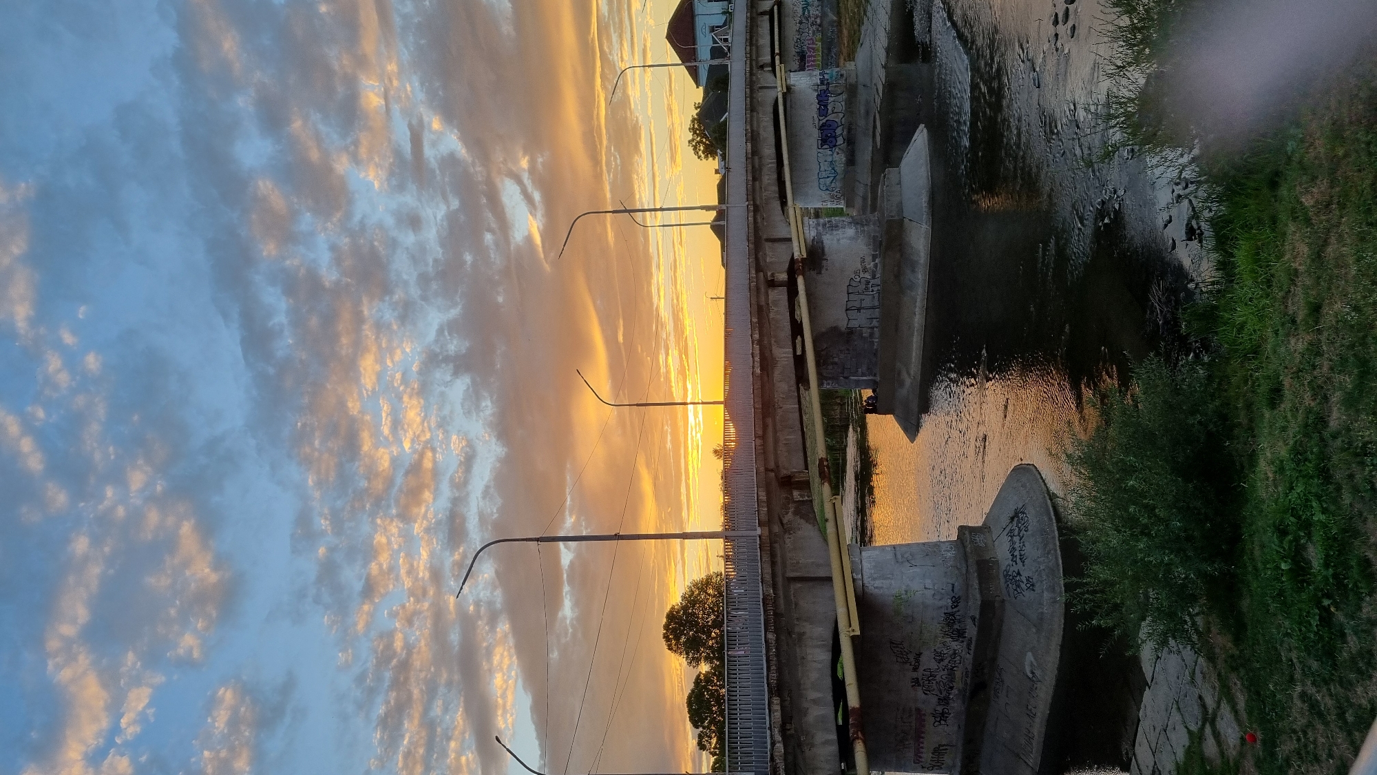 The image captures a sunset over a bridge in Mukachevo, Ukraine. The sky is filled with colorful clouds, transitioning from blue to orange and yellow hues. The bridge, constructed of concrete and metal, spans a calm river. The bridge's concrete pillars are covered in graffiti. Streetlights are visible along the top of the bridge. Trees line the far bank of the river, and a few buildings can be seen in the distance. The foreground shows a grassy embankment leading down to the water's edge. The scene suggests a peaceful evening in an urban setting.