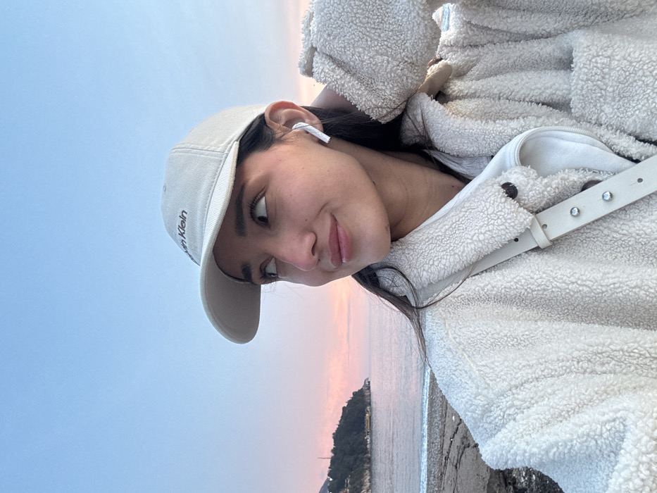 A medium close-up, portrait-oriented image shows a young woman captured in an outdoor coastal setting, likely in Budva, Montenegro. The photo is rotated 90 degrees clockwise.
The main subject is a young woman with dark hair, fair skin, and a serene expression, looking towards the right side of the frame. She wears a light beige baseball cap, on which the word "Klein" is partially visible, indicating "Calvin Klein". She has a white AirPod in her right ear and is dressed in a cozy, cream-colored sherpa or fleece jacket. A light-colored bag strap with metallic studs is visible over her shoulder.
The background features a tranquil seascape. The sky displays a beautiful gradient of colors, transitioning from light blue at the top to soft pink and orange hues closer to the horizon, strongly suggesting either sunrise or sunset. A calm body of water, likely the sea, stretches into the distance, with a prominent landmass or small island featuring structures visible on the far side. The foreground also shows a hint of a beach or rocky shoreline.
The woman appears to be posing for a photograph, possibly a selfie, enjoying the coastal ambiance during the golden hour. Her gaze is gentle, conveying a sense of peace. The soft natural lighting enhances the warm, relaxed mood of the scene.