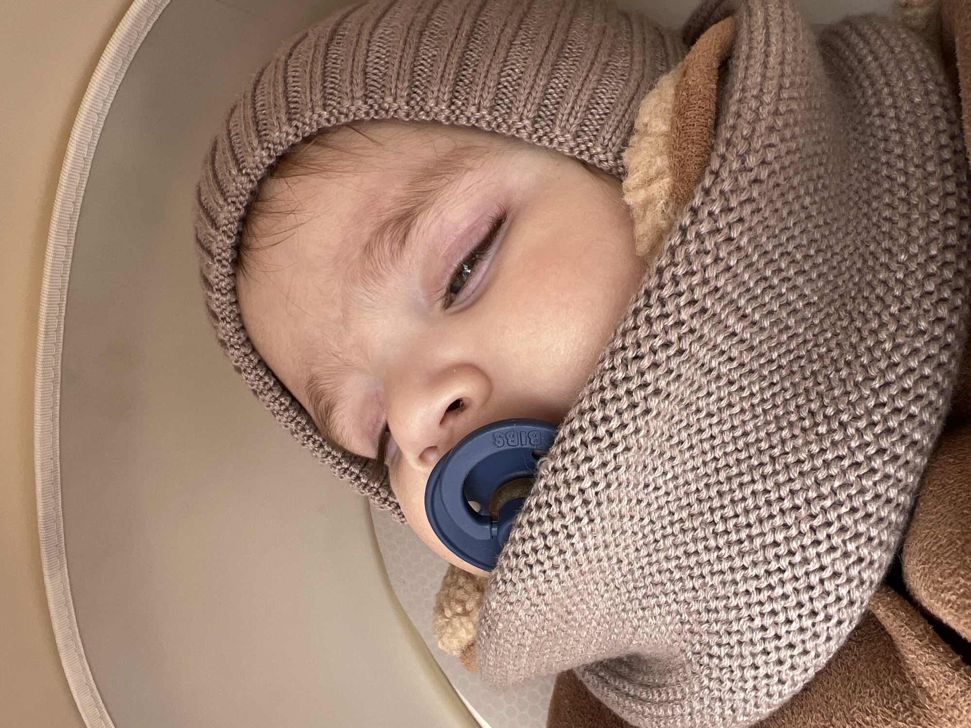 A baby lies with their eyes mostly closed, wearing a light brown knit beanie and a larger knit scarf or blanket in a similar tone. The baby has a blue pacifier in their mouth. The background is a beige color. The word "BIBS" is visible on the pacifier. The location and city are not discernible from the image.