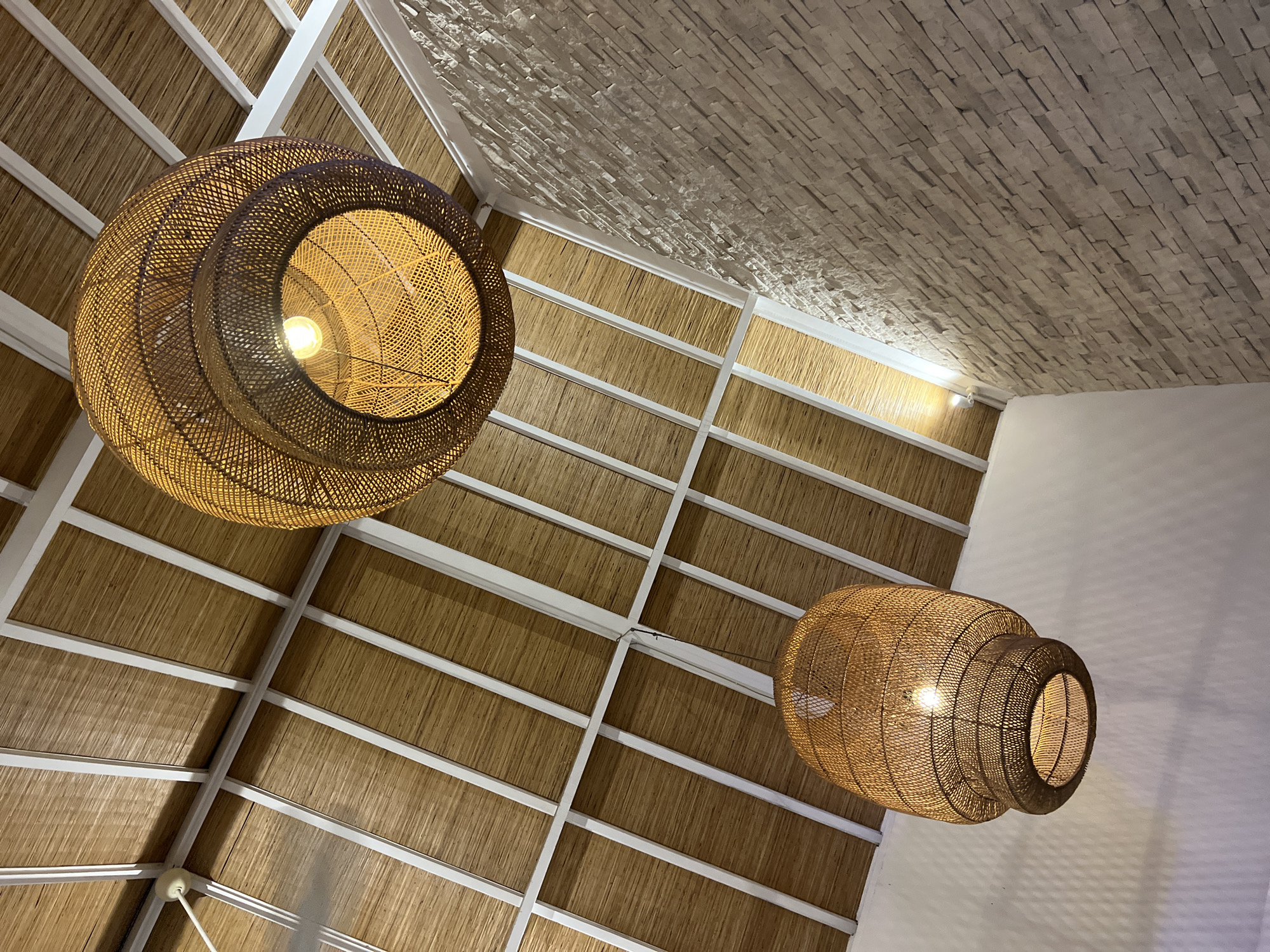 The image captures an interior view, looking upwards towards a ceiling. Two large, intricately woven, rattan lamps are suspended from the ceiling. The lamp on the left is spherical, while the lamp on the right is more vase-shaped. Both lamps are illuminated, casting a warm, amber glow. The ceiling structure consists of a white grid with a woven, possibly bamboo, material filling the spaces between the grid. A section of the ceiling is constructed from light-colored, stacked, rectangular stones or tiles. The walls are painted white. The scene appears to be in a building in North Kuta, Indonesia. No people are visible in the image.